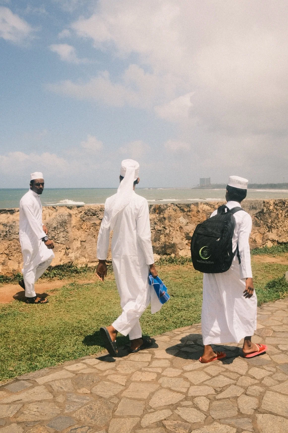 Three men dressed in white traditional clothing walking near the coast with a stone wall, ocean, and partly cloudy sky in the background.