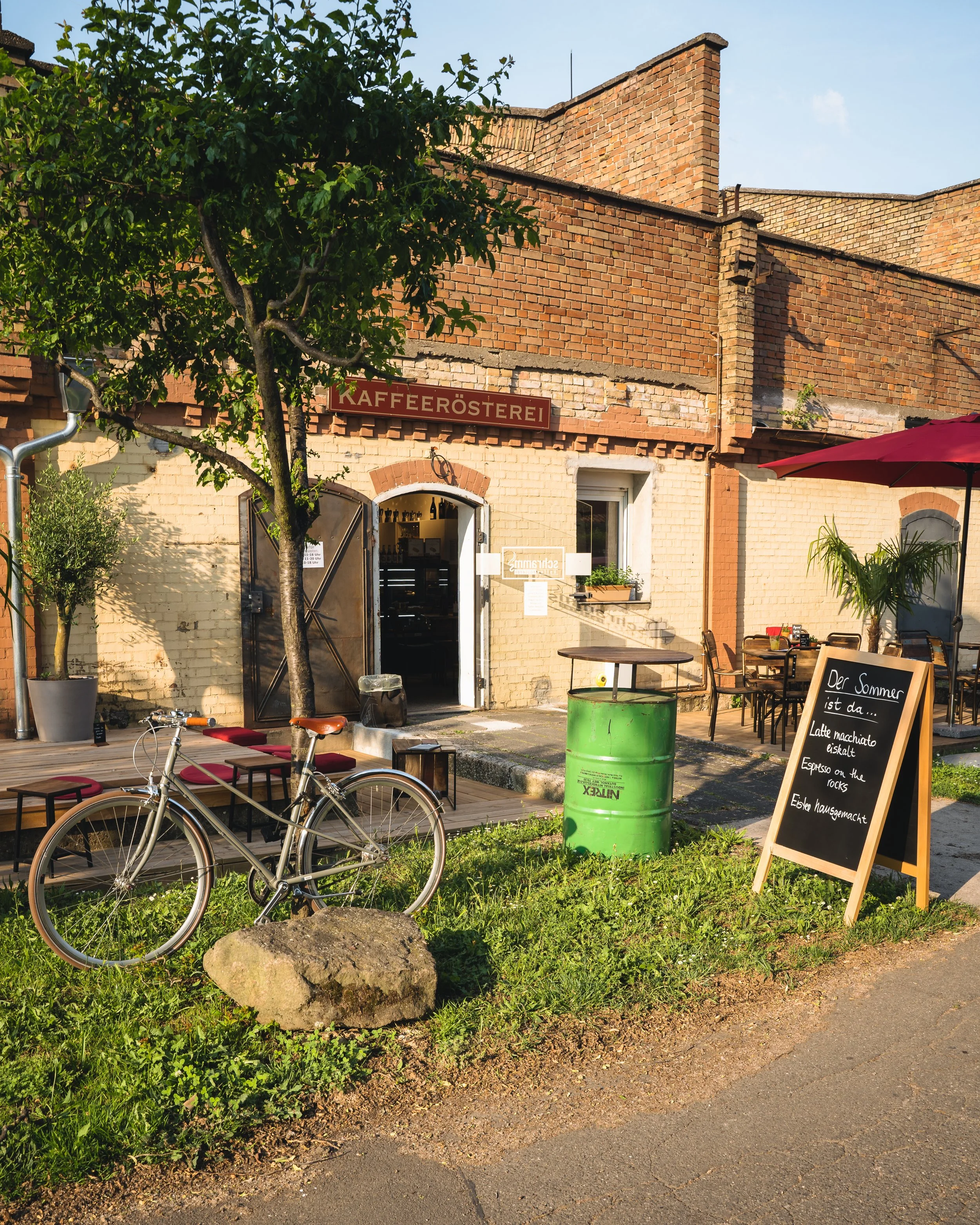 Outdoor scene of a cafe with a brick building, a tree, and outdoor seating. There's a blackboard sign with menu items, and a green barrel used as a table. A bicycle is parked in front of the cafe.