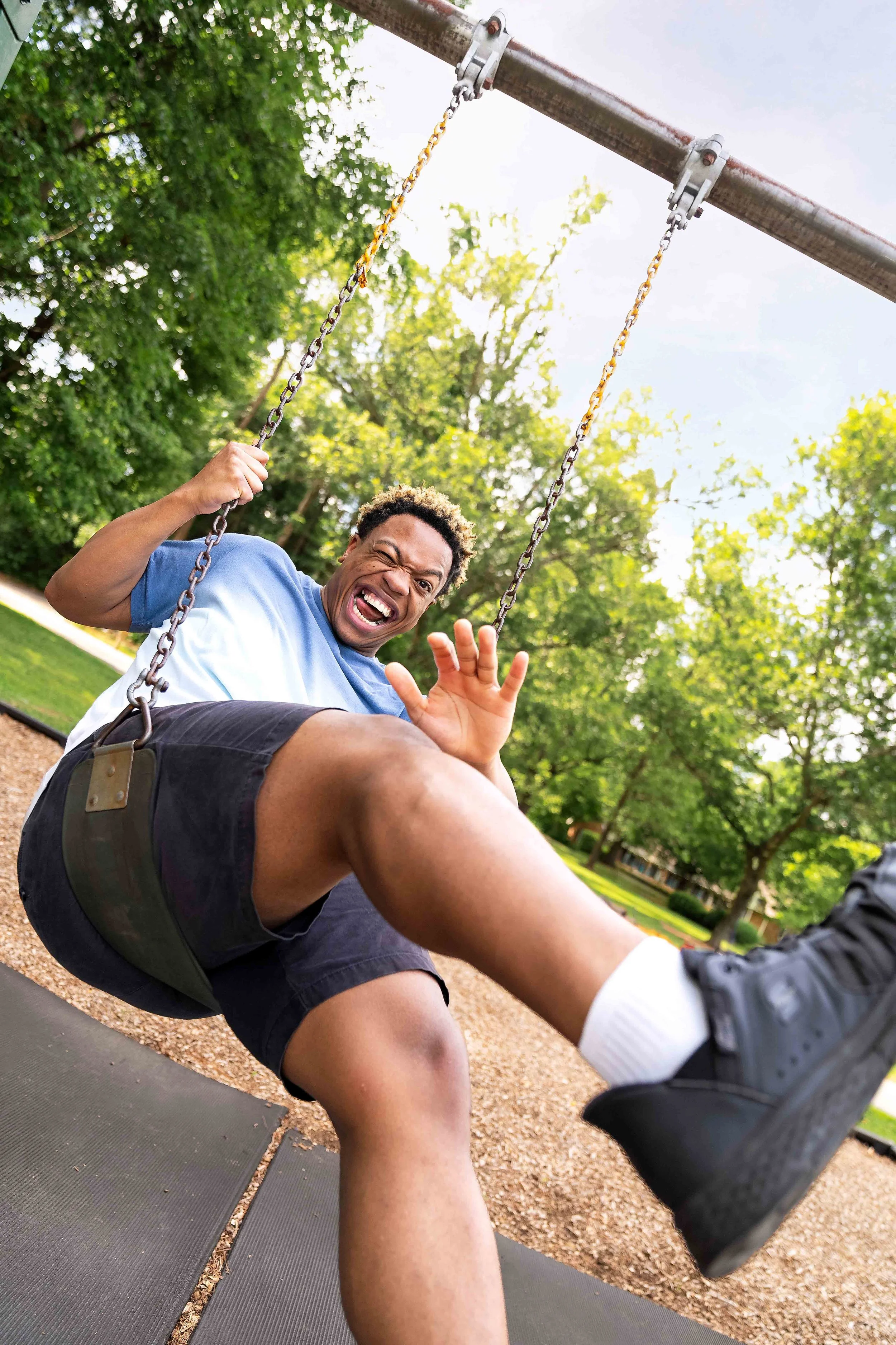 A man on a swing at a park, smiling and making a humorous face, wearing a blue t-shirt and black shorts, with trees in the background.
