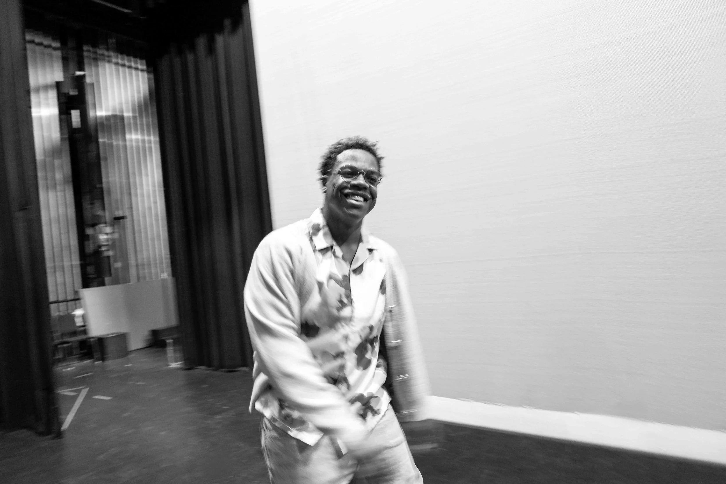 A smiling man with glasses wearing a patterned shirt standing indoors next to an elevator, with a plain wall behind him.