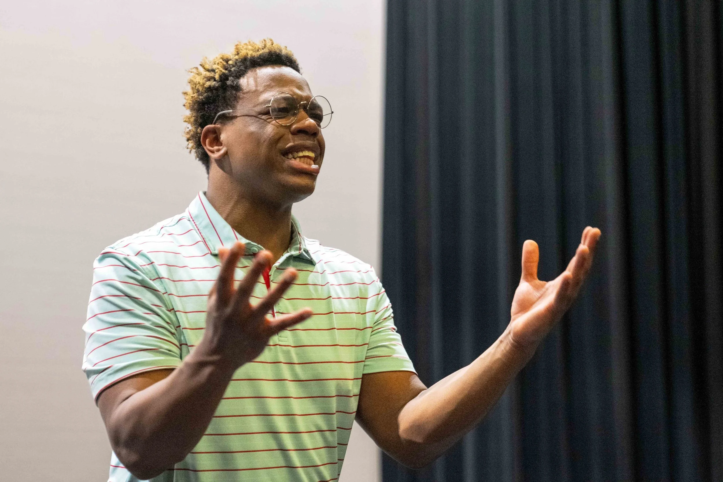 A person wearing glasses and a striped shirt, gesturing with hands, speaking in front of a dark curtain.