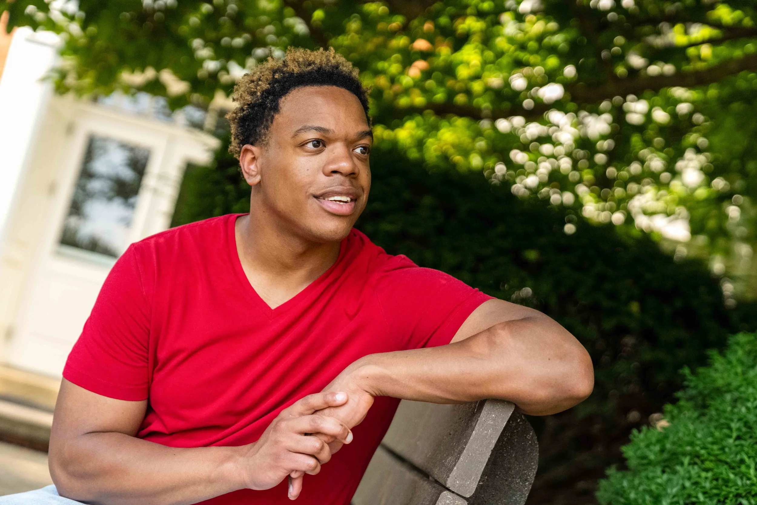 A man in a red t-shirt sitting on a park bench outdoors, looking to the left with a neutral expression, surrounded by greenery and trees in the background.