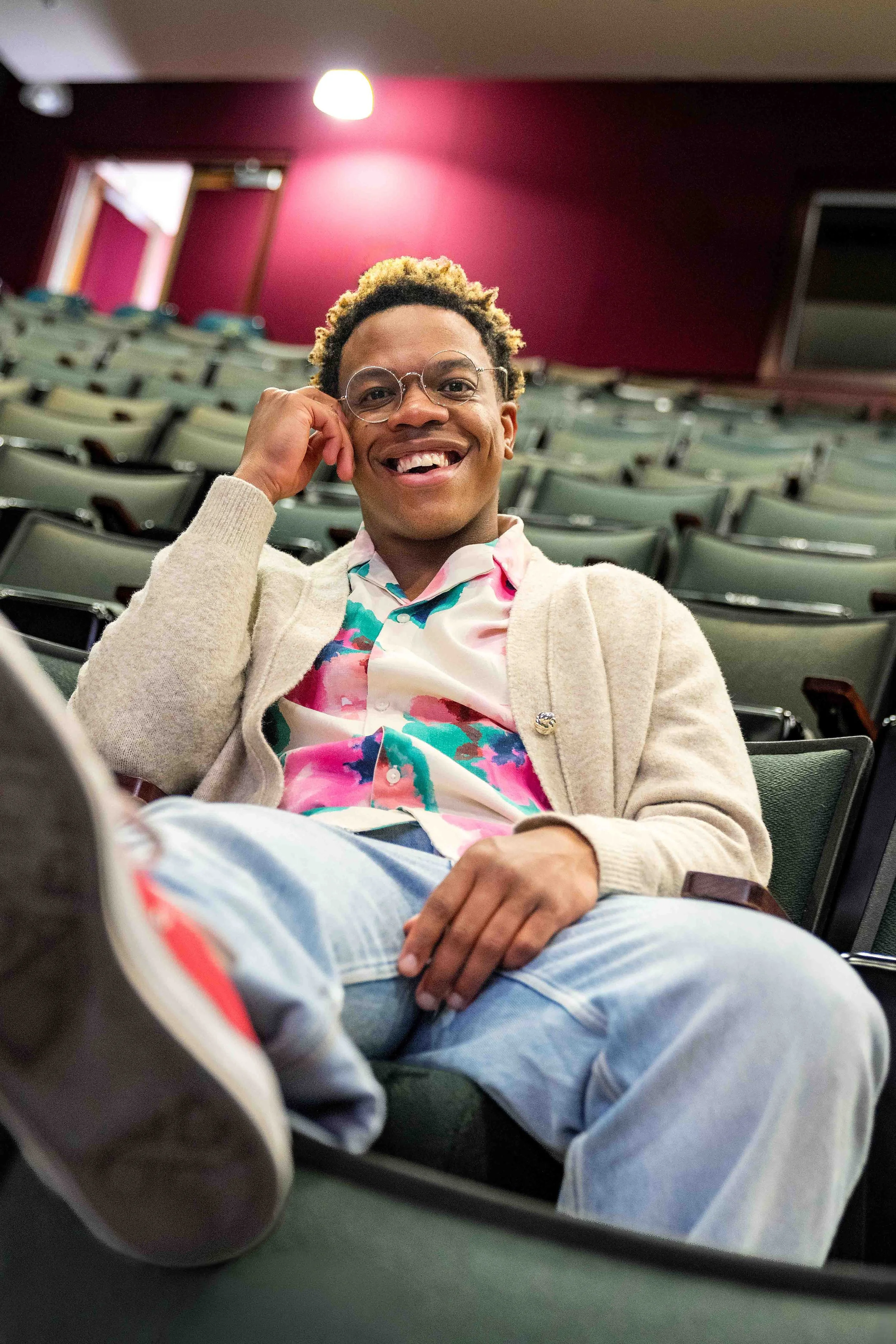 Young man with curly blonde hair and glasses smiling while sitting in an auditorium with red walls and green chairs, with a bright light above him.