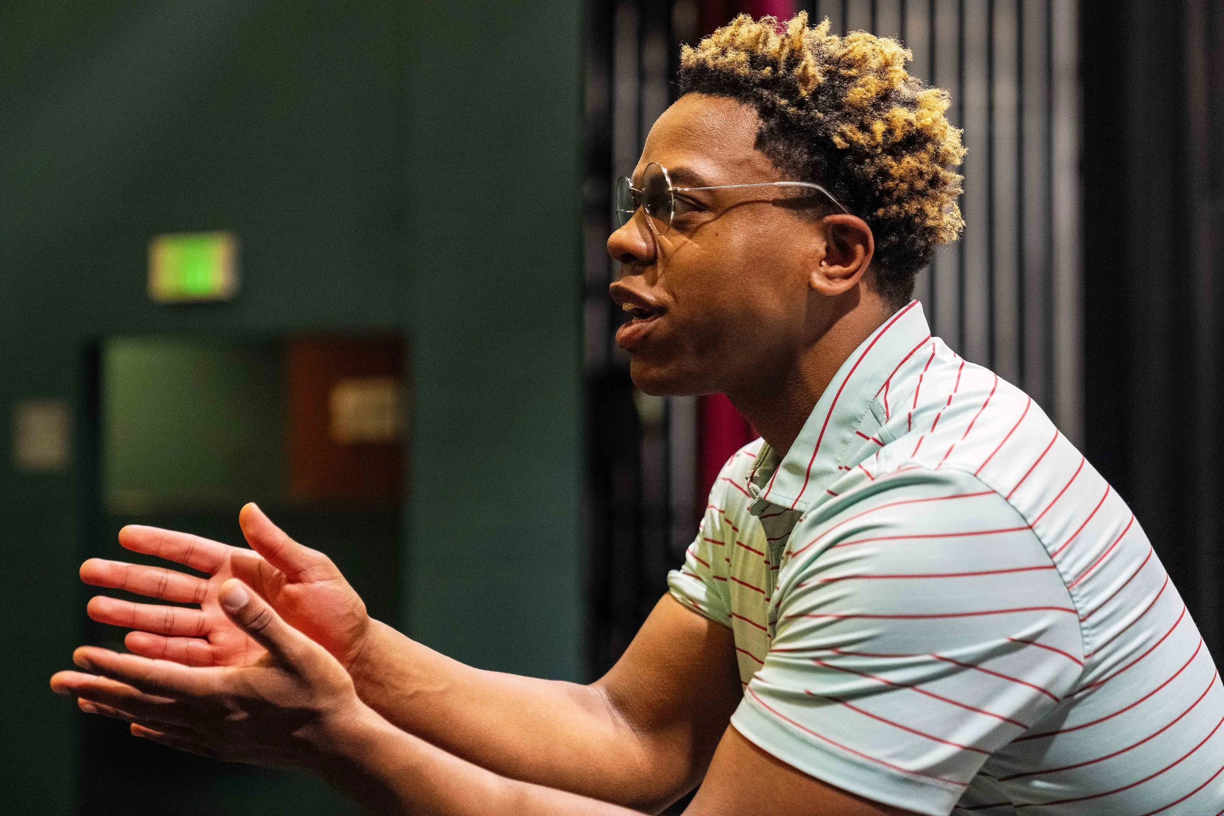 A person with light brown skin, short curly hair, sunglasses, and a striped shirt gesturing with their hands while speaking.
