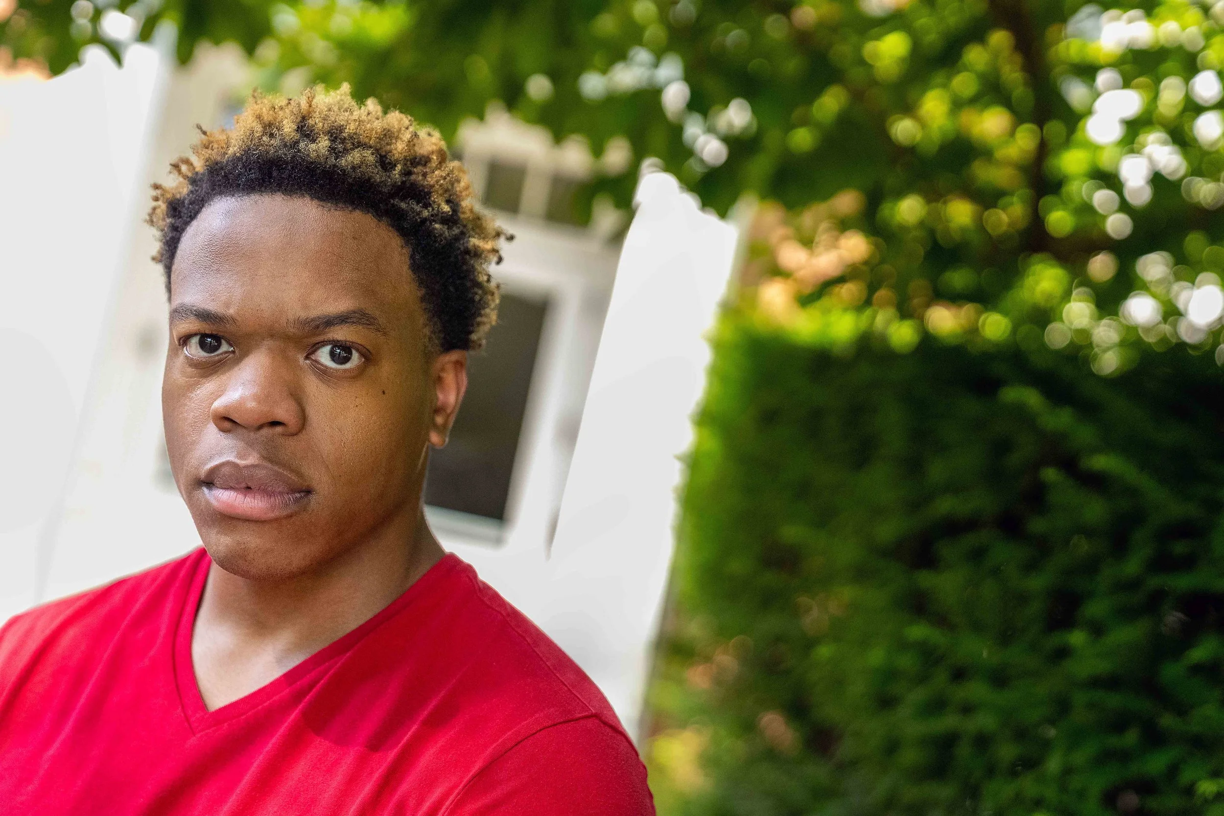 A young man with curly, blonde-tipped hair wearing a red shirt outdoors with green trees in the background.