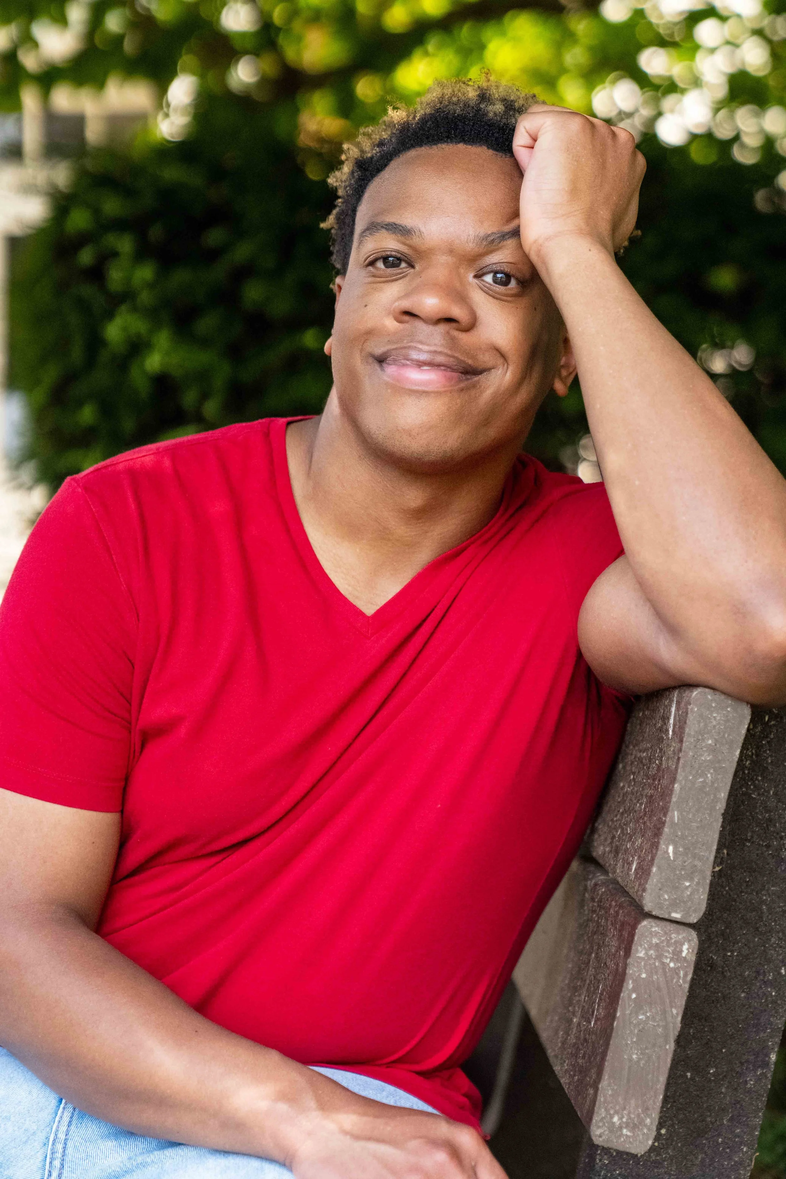 A young person with short curly hair, wearing a red shirt, smiling, resting their head on their hand while sitting on a park bench with greenery in the background.