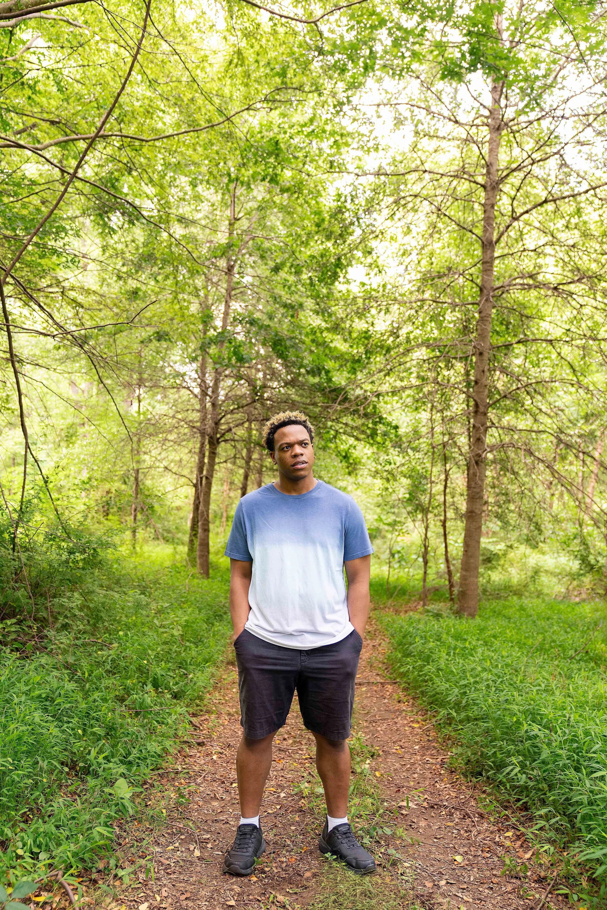 A young man stands on a dirt trail in a lush green forest, with trees and foliage surrounding him.
