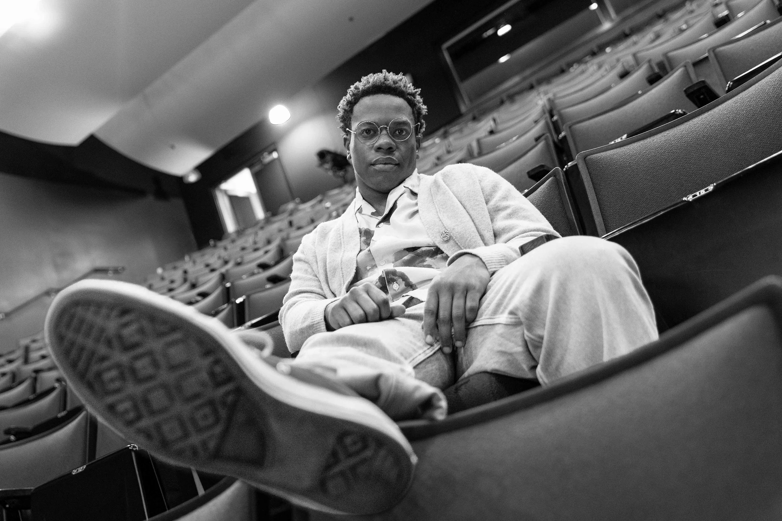 A young man with curly hair and glasses sitting in an empty auditorium, viewed from a low angle.
