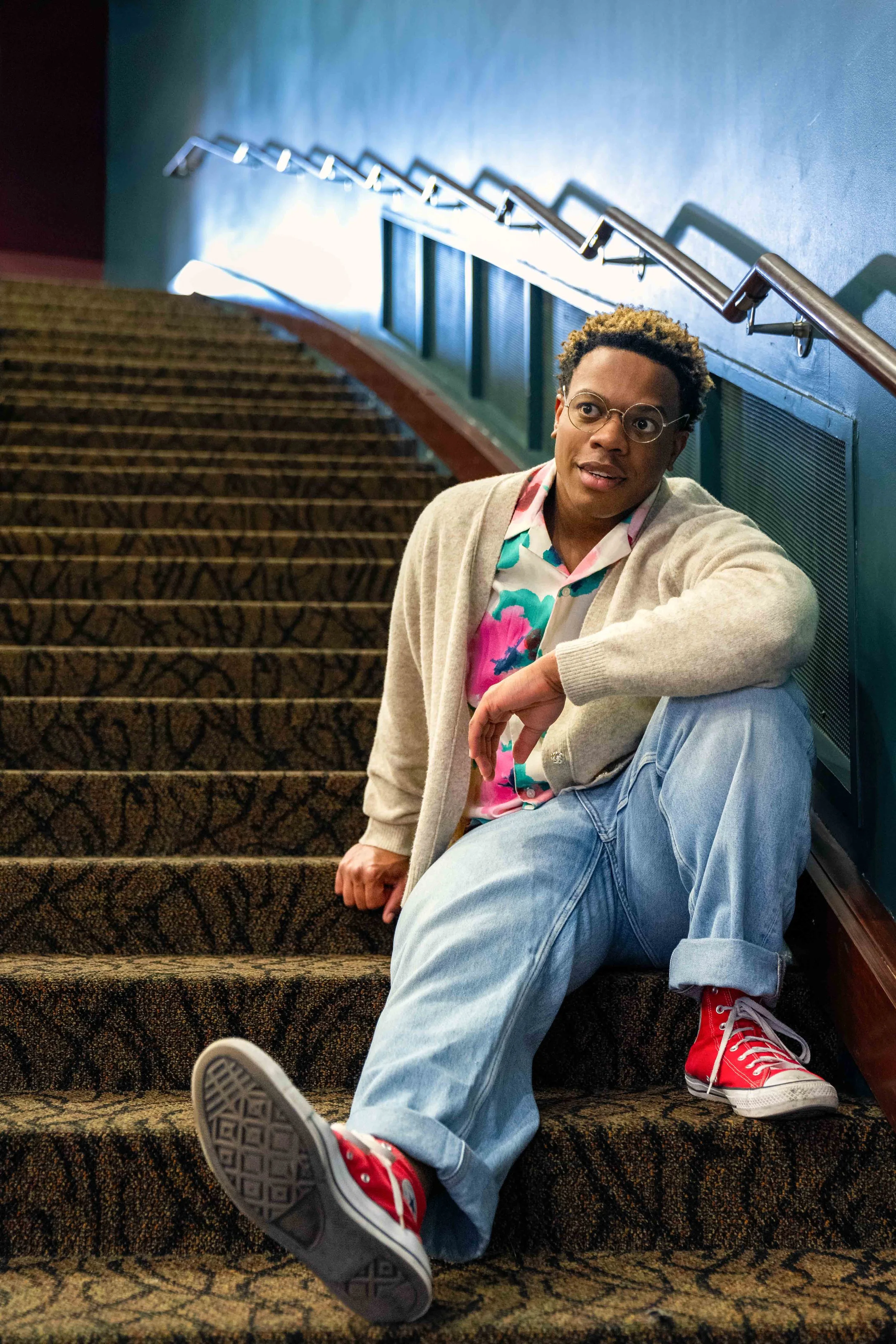 A young person with curly hair, glasses, and colorful clothing sitting on stairs made of patterned carpet, leaning against the wall with a relaxed expression.