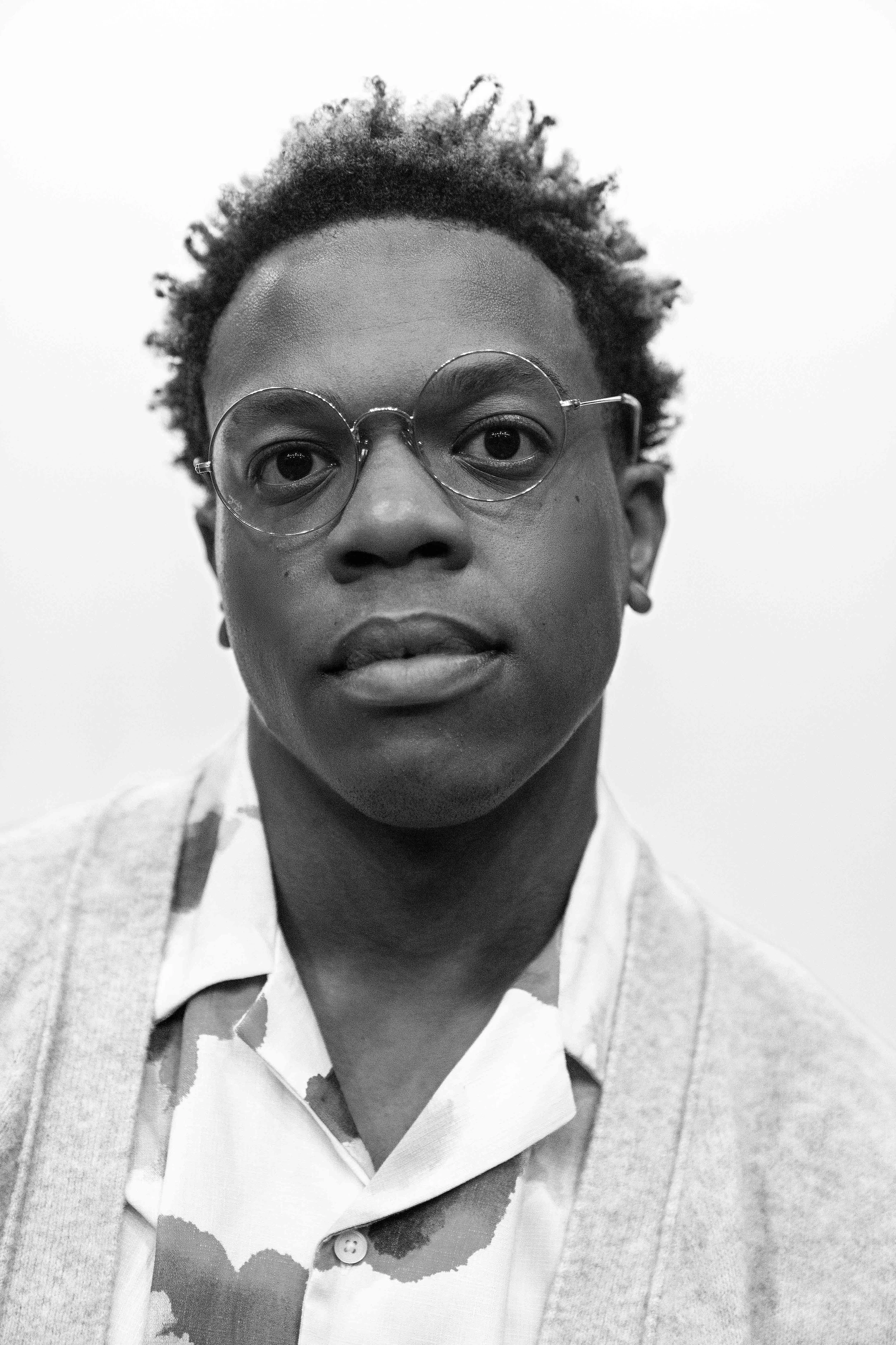 Black and white portrait of a man wearing round glasses, a white collared shirt, and a light sweater, looking directly at the camera.