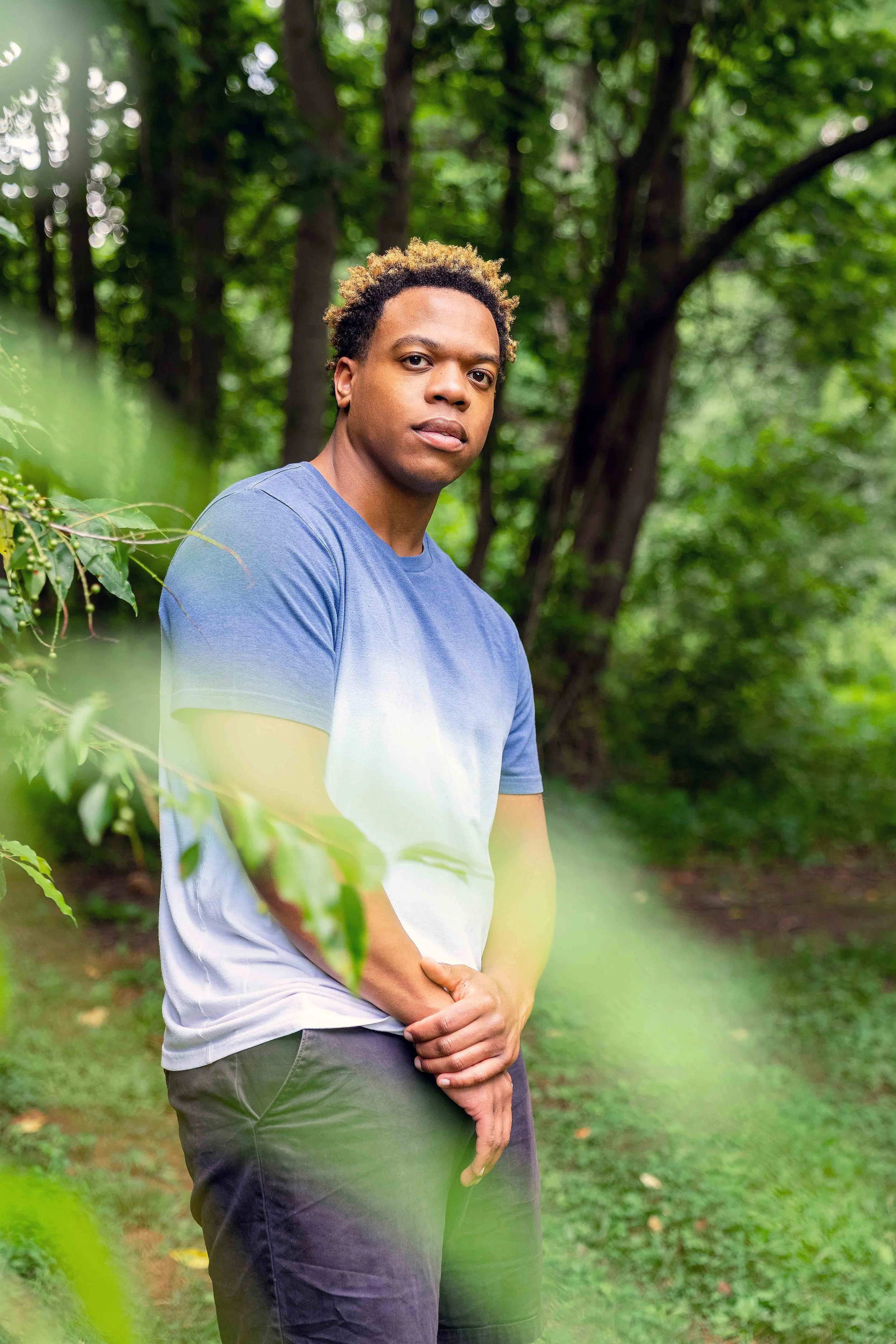A young man with curly blond-tipped hair wearing a blue and white t-shirt and dark pants standing outdoors amid green trees and foliage, looking directly at the camera with a neutral expression.