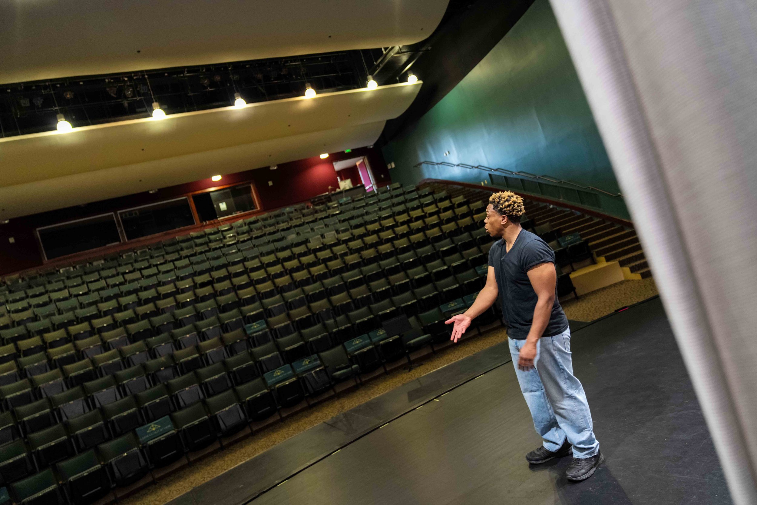 A man standing on stage in an empty auditorium, gesturing with his hands, with rows of seats and a balcony in the background.