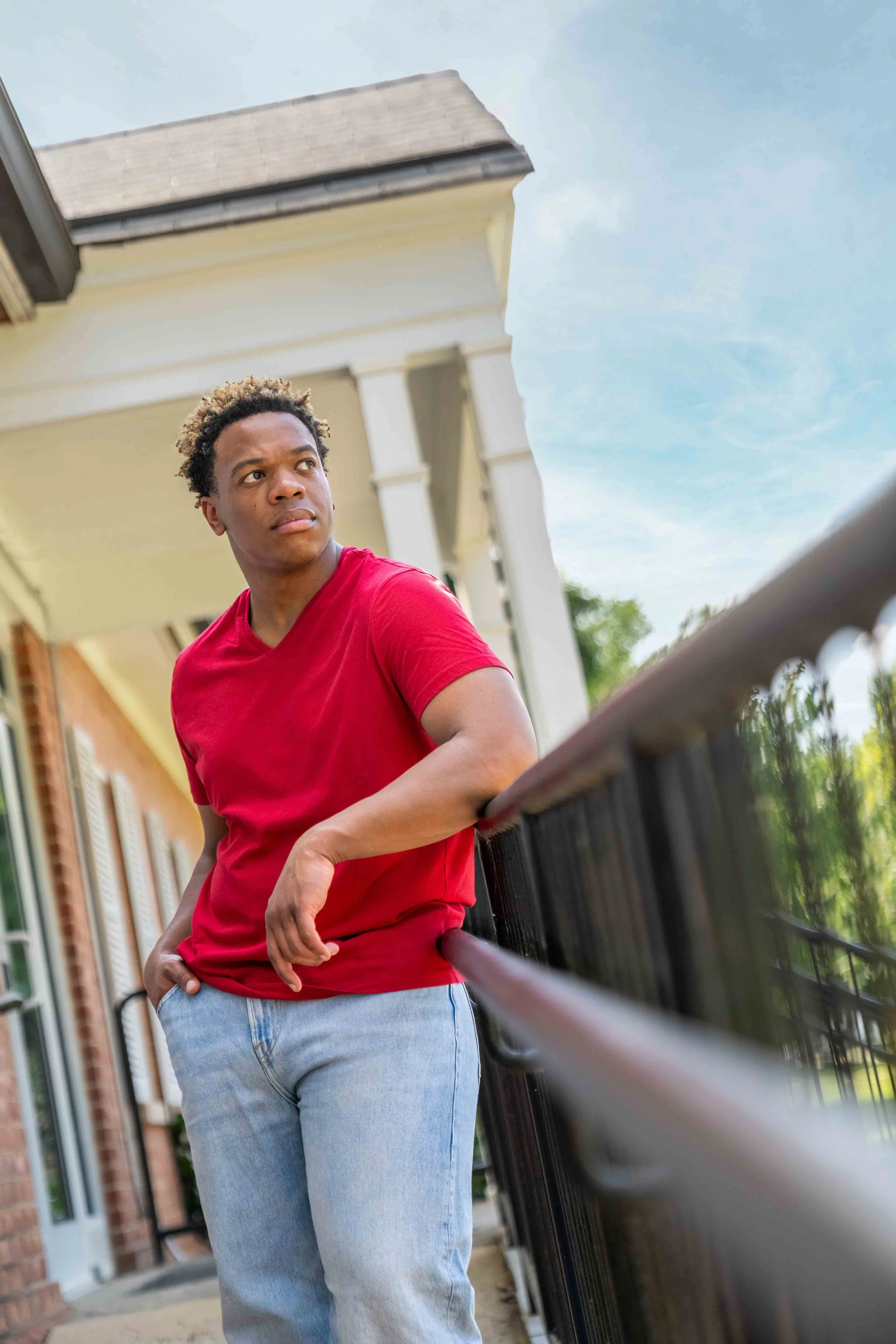 A young man with curly hair wearing a red T-shirt and light blue jeans standing on a porch near a black railing, with a house and blue sky in the background.
