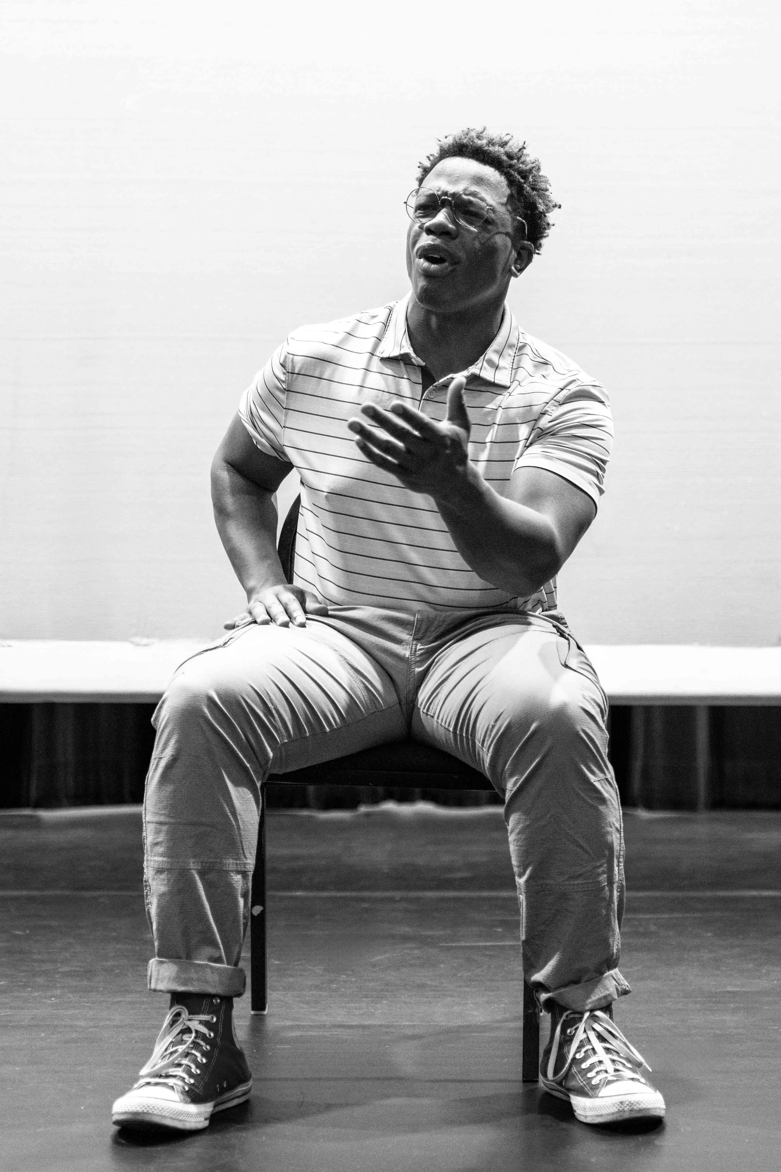 A young man sitting on a chair on a stage, wearing glasses, a striped polo shirt, rolled-up pants, and sneakers, with one hand on his chest and the other on his thigh, speaking passionately.