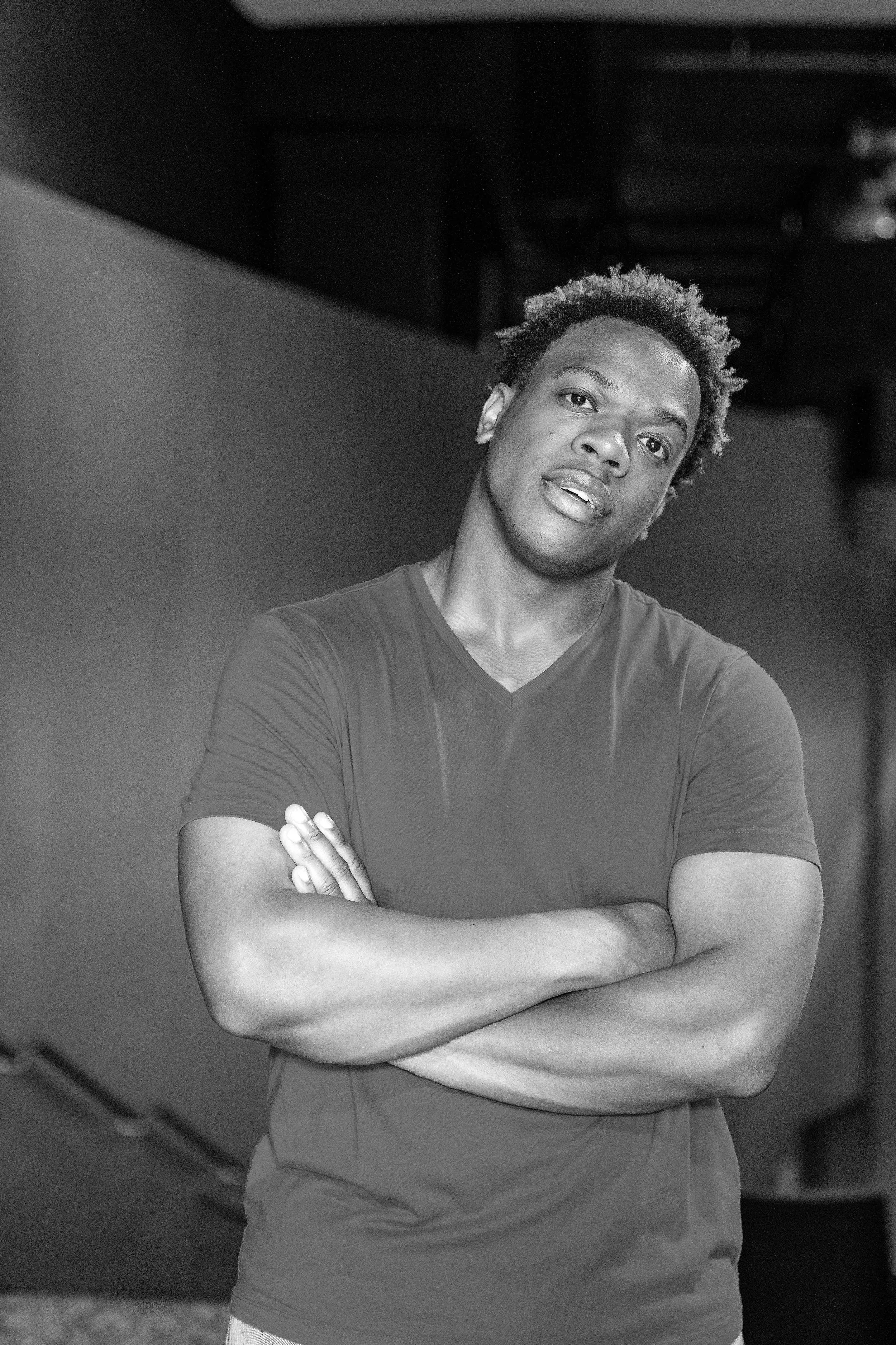 A black and white photo of a man with short curly hair standing with arms crossed, looking at the camera. He is wearing a short-sleeved t-shirt and appears to be in an indoor setting.