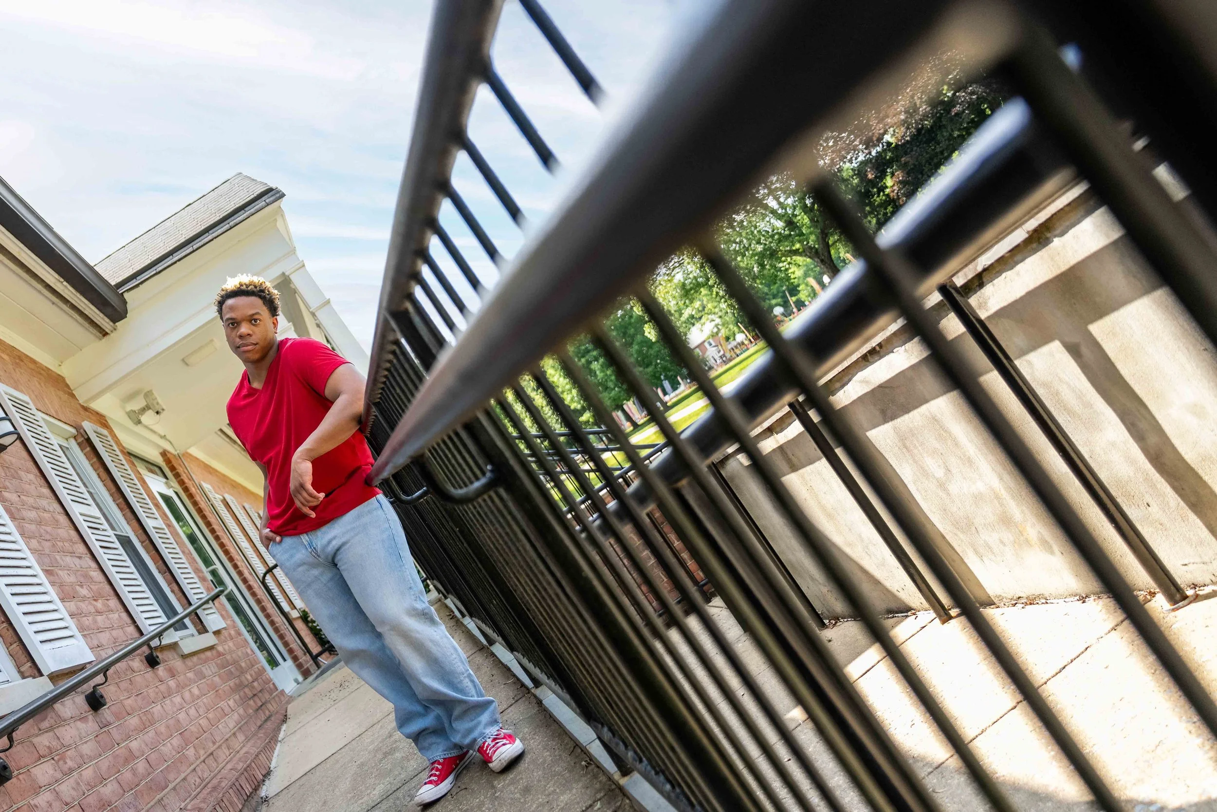 Young man in red t-shirt and jeans standing behind a metal railing in front of a brick house with white window shutters, looking directly at the camera in an outdoor neighborhood setting.
