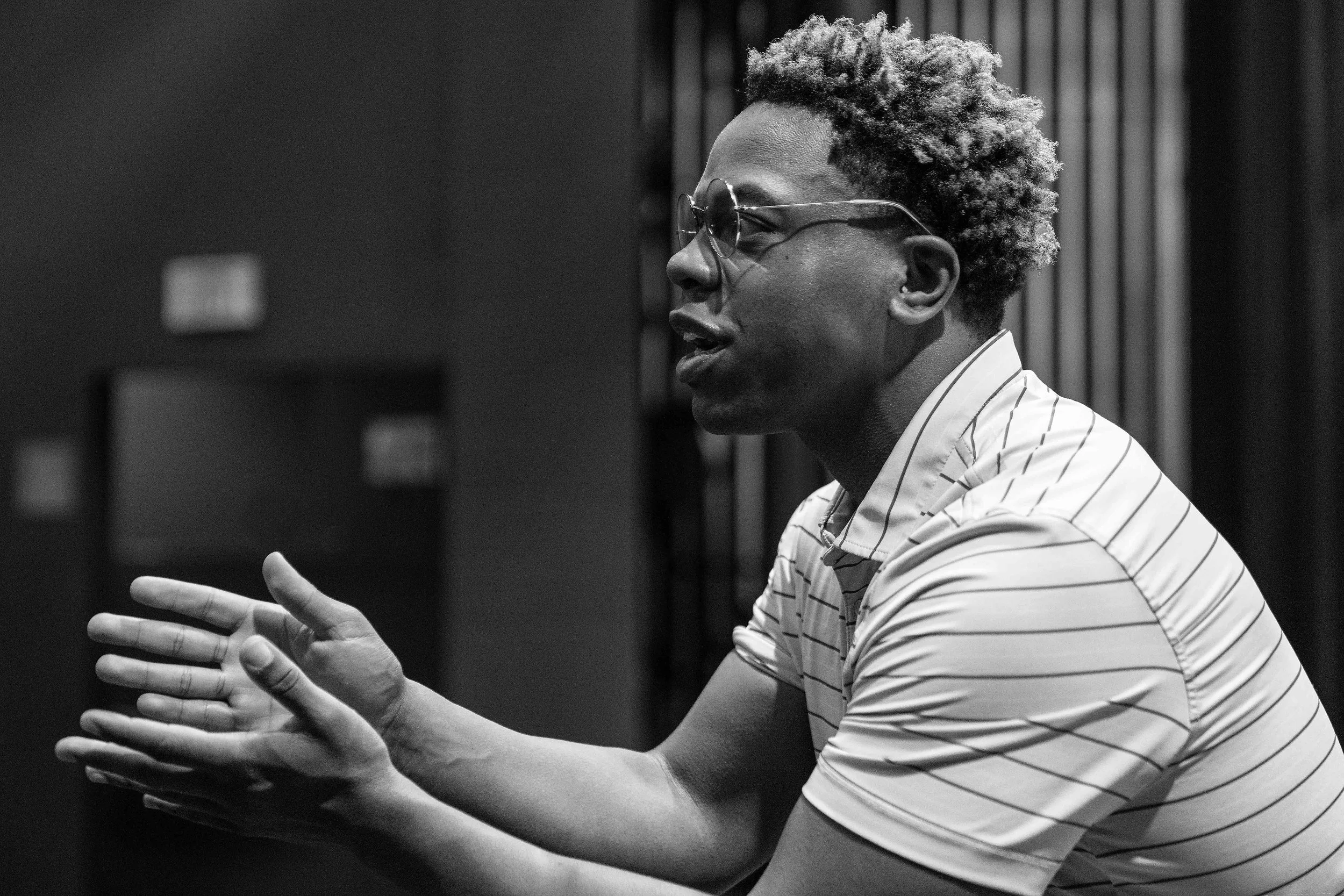 A black and white photo of a man with glasses and curly hair talking or explaining, with his hands gesturing, in a professional or casual setting.