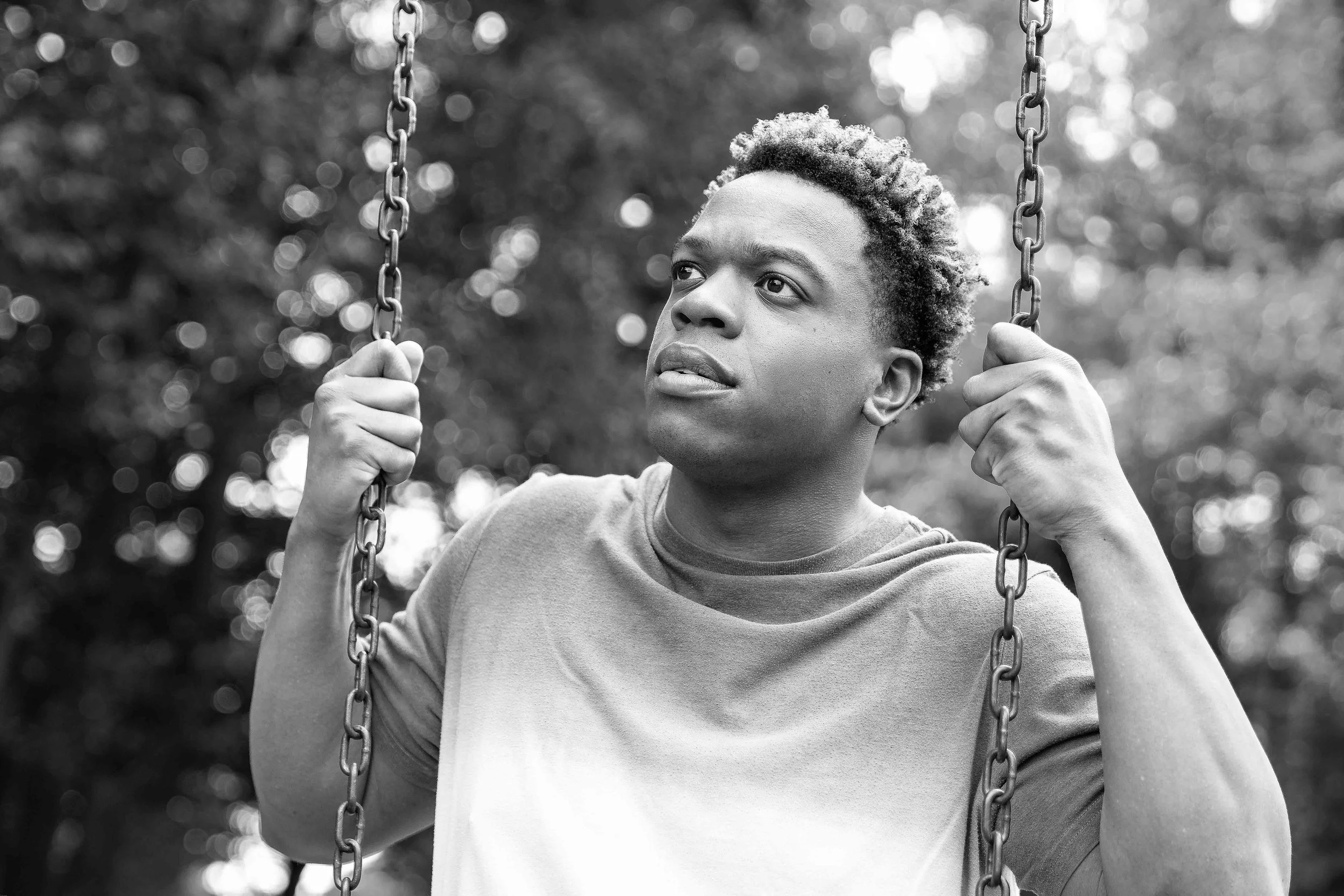 A young person with short, curly hair sitting on a swing at a park, looking into the distance with a thoughtful expression, black and white photo.