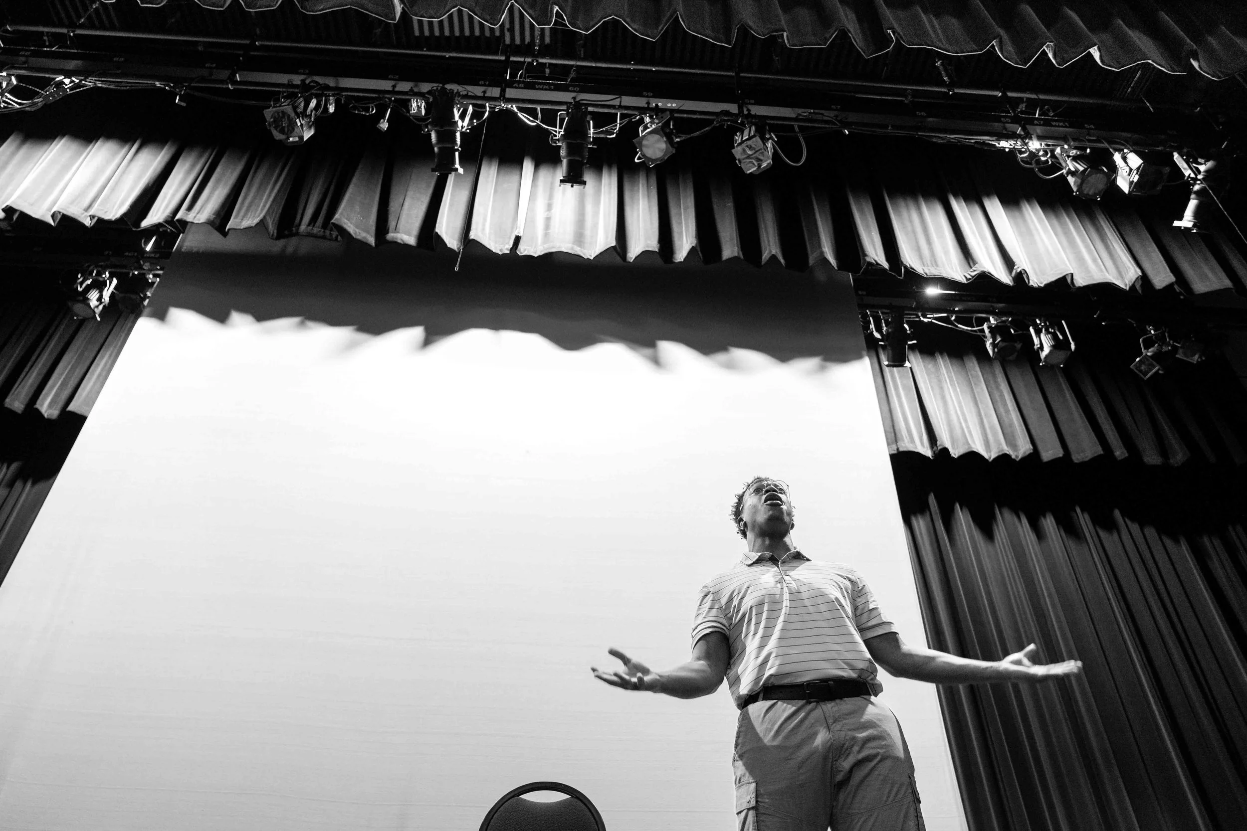 A man standing on a stage with curtains, gesturing with his arms, during a presentation or performance in a theater or auditorium.