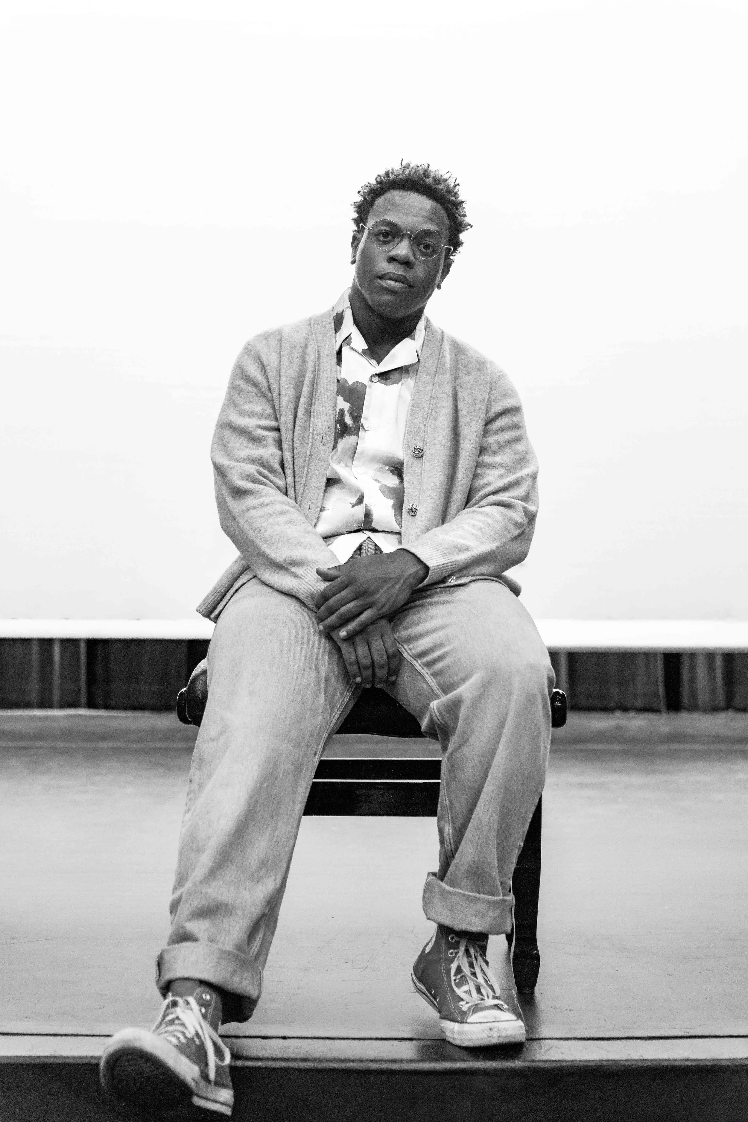 A young man with curly hair and glasses sitting on a chair in a room with a plain white wall. He is wearing a patterned shirt, a cardigan, and loose jeans with sneakers. The photo is in black and white.