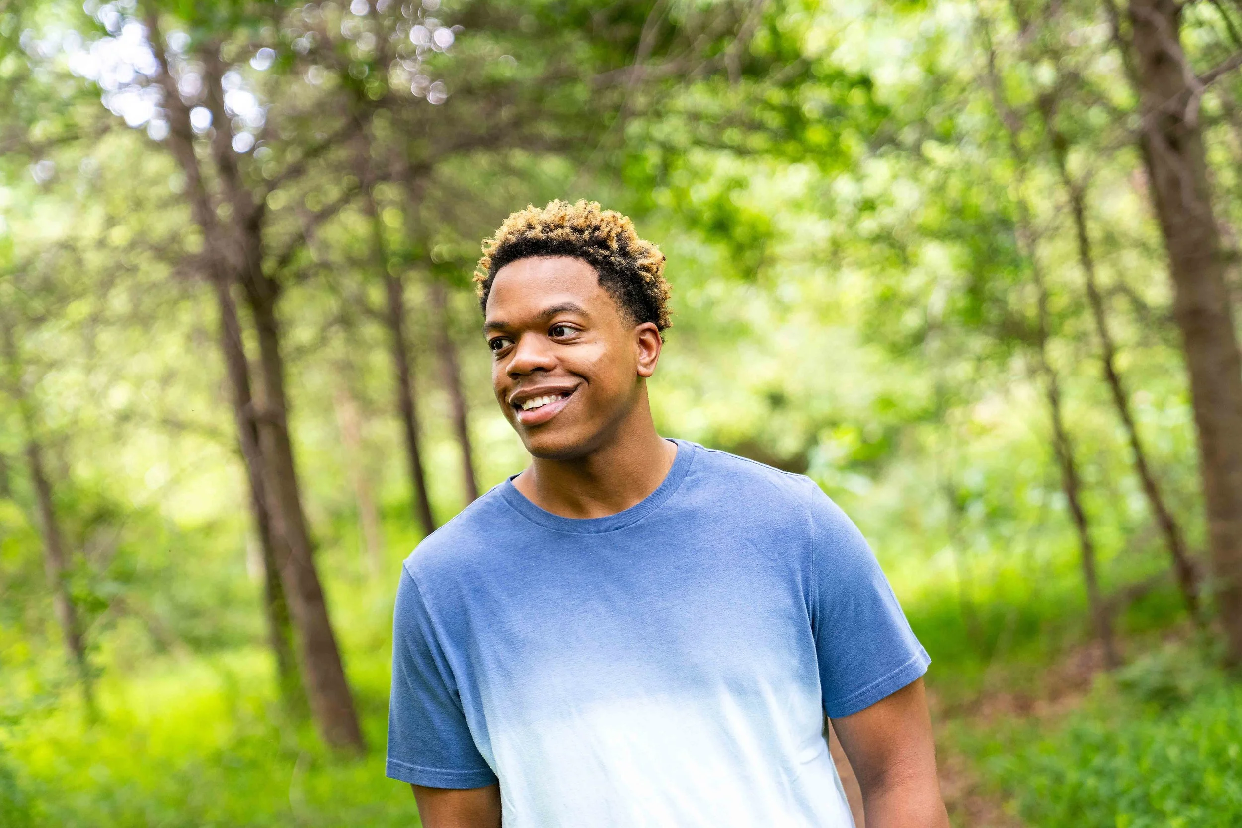 A young man with short, curly hair with blond tips smiling and looking to his right in a lush, green forest.