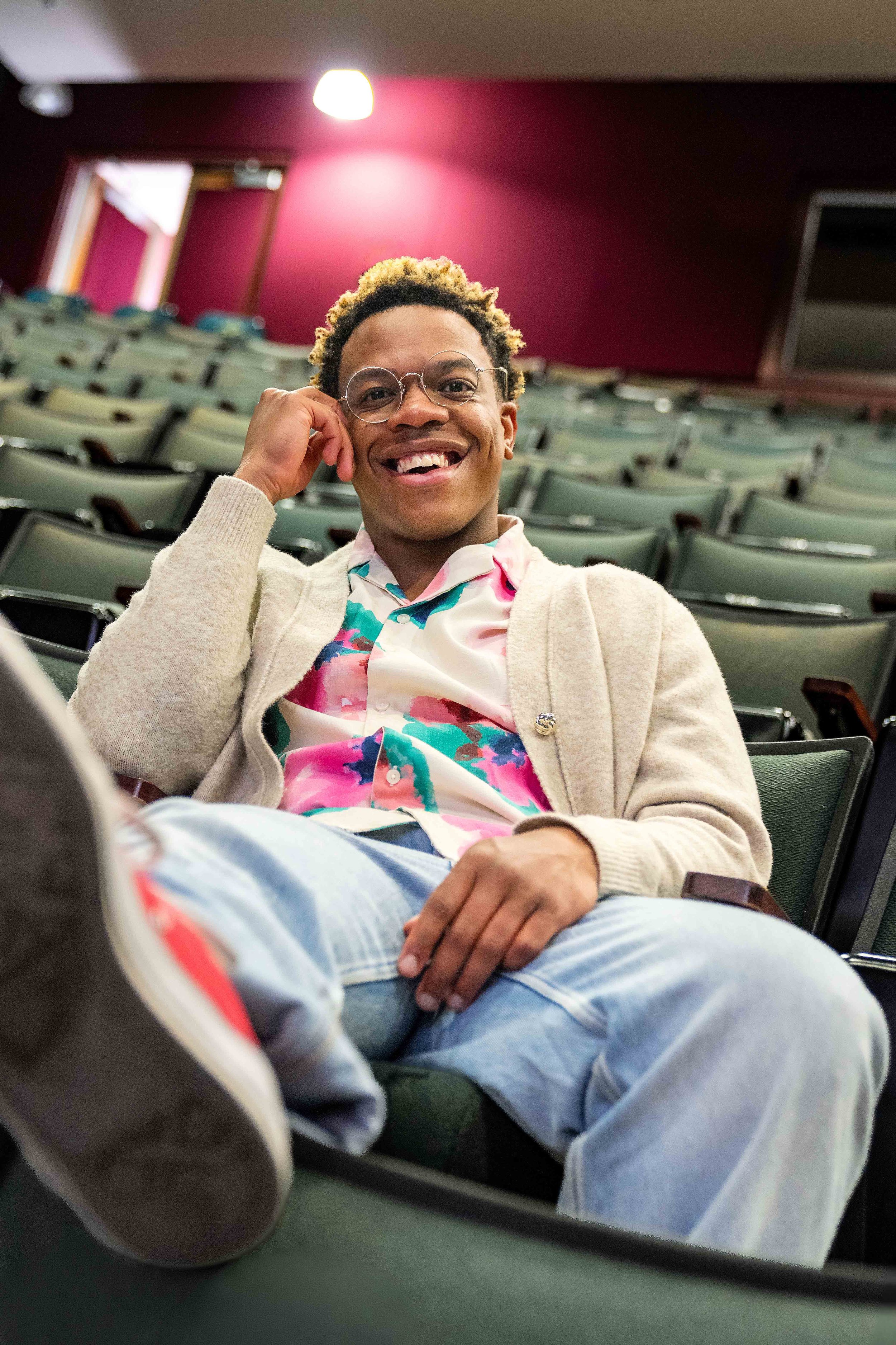 A young man with blond highlights in his hair, wearing glasses, a colorful shirt, a beige cardigan, and jeans, sitting in an empty auditorium or classroom, smiling and looking at the camera.