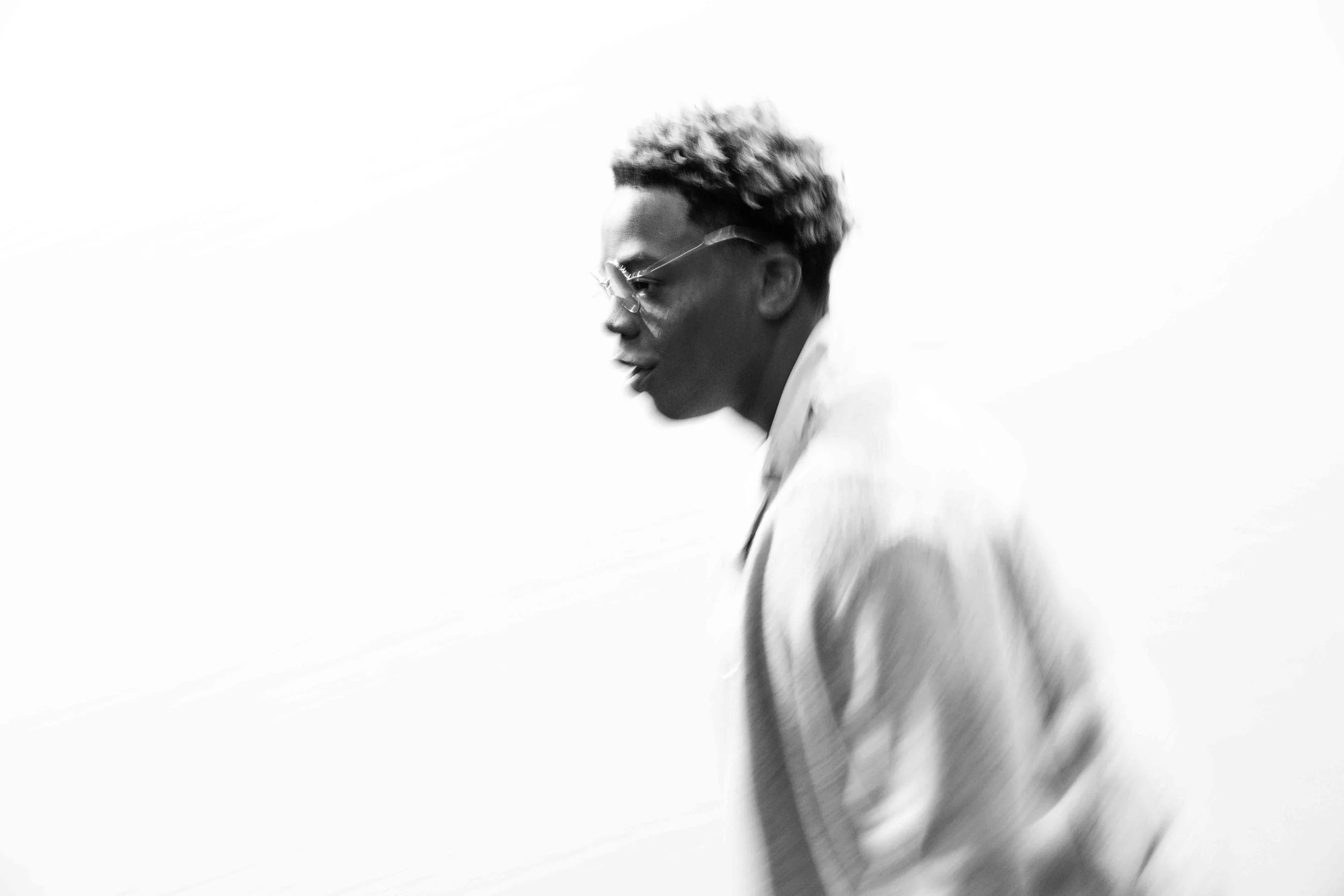 A black and white photograph of a man with short, curly hair wearing glasses and a white top, captured in profile with a bright, minimal background.