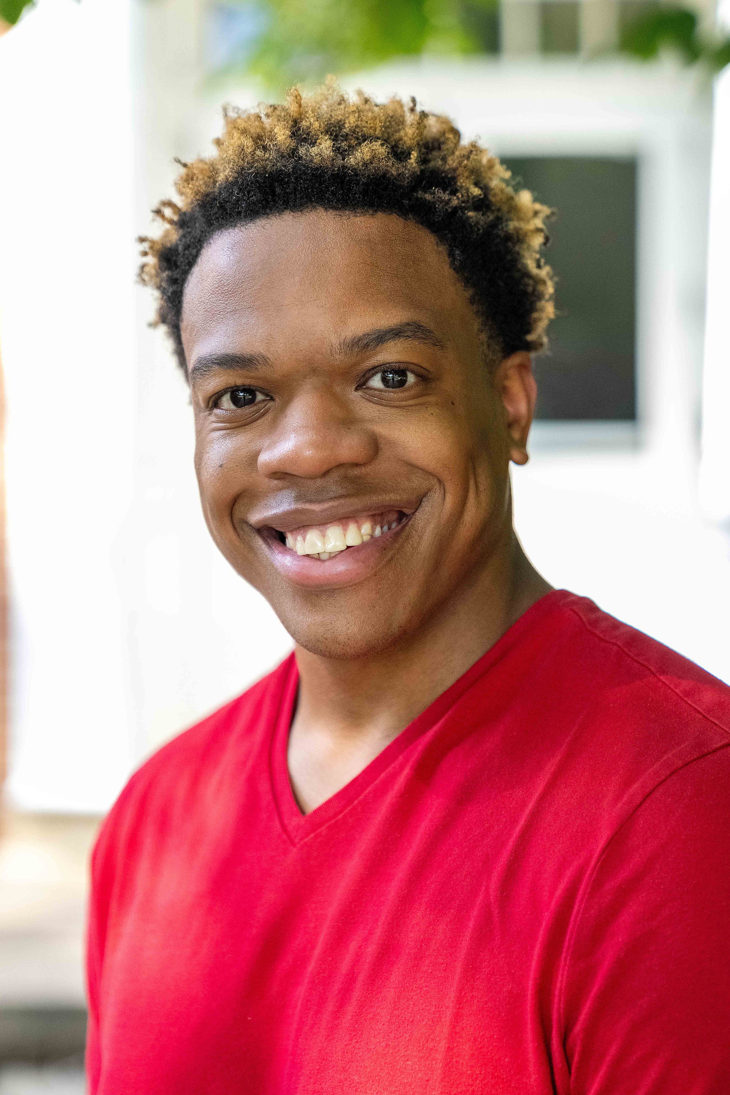 Close-up of a smiling young man with short, curly hair, wearing a red shirt, indoors with a blurred background.