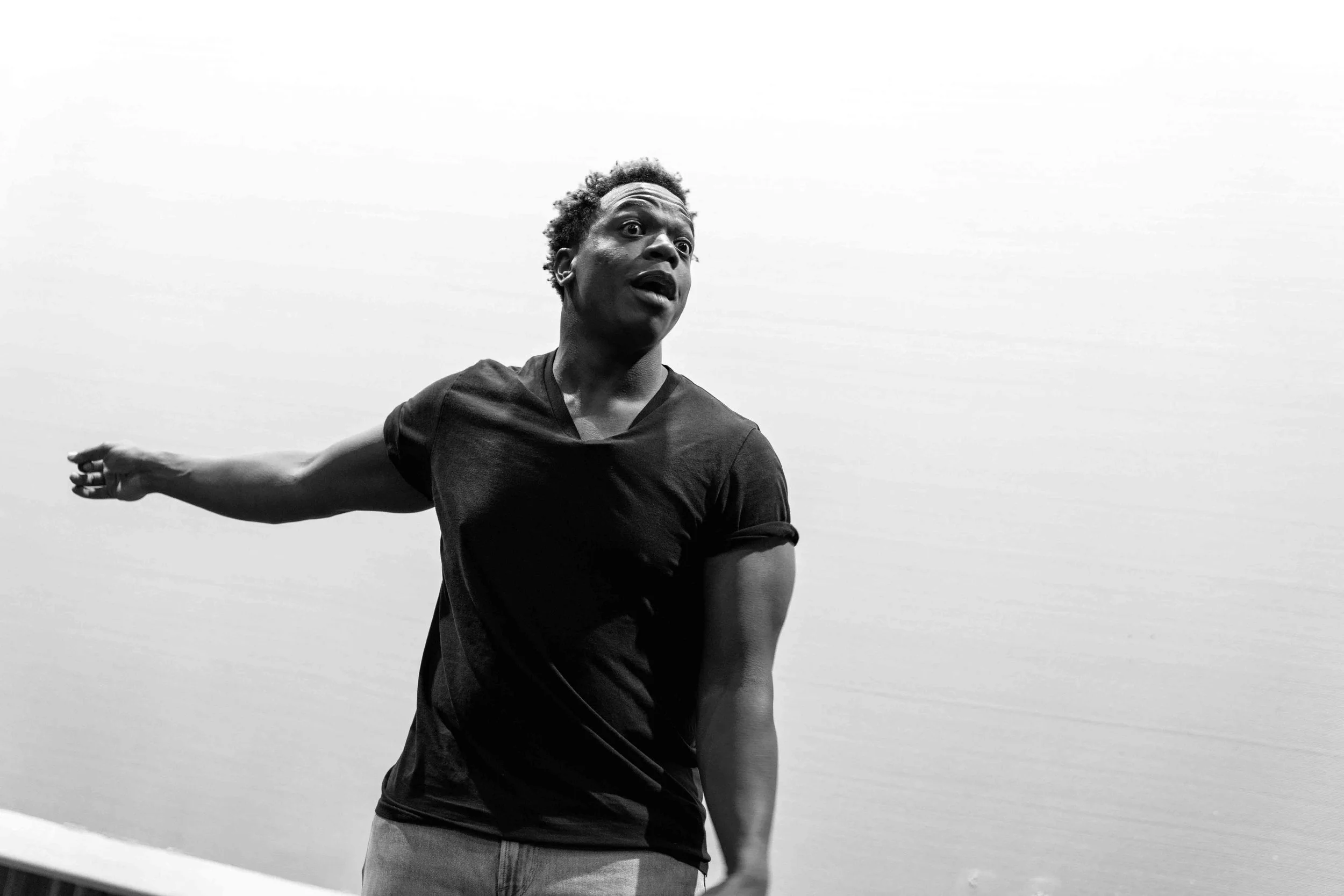 Black and white photo of a person with short curly hair in a dark shirt, reaching out with one arm, outdoors against a cloudy sky.