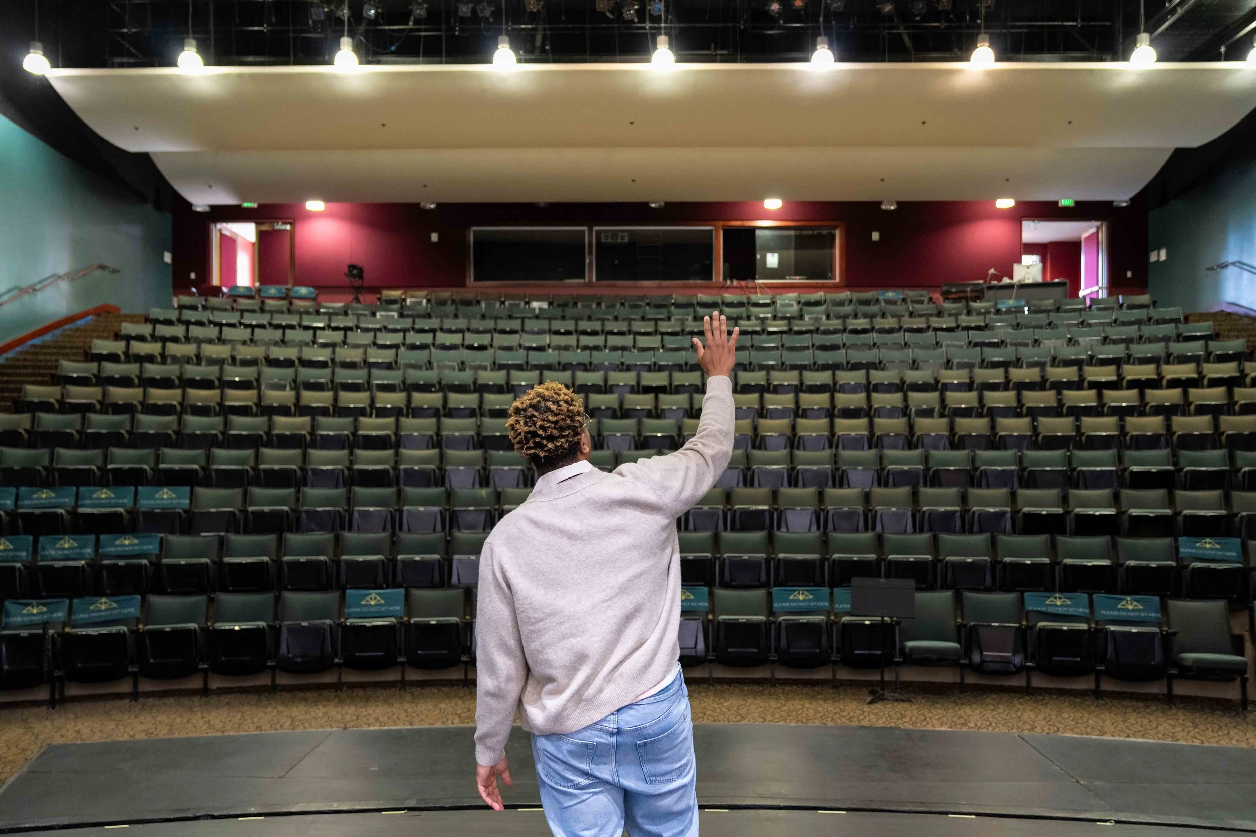 A person with curly hair wearing a light gray sweater and blue jeans standing on a stage in an empty auditorium, raising their right arm with a hand open, facing towards rows of green seats and the stage.