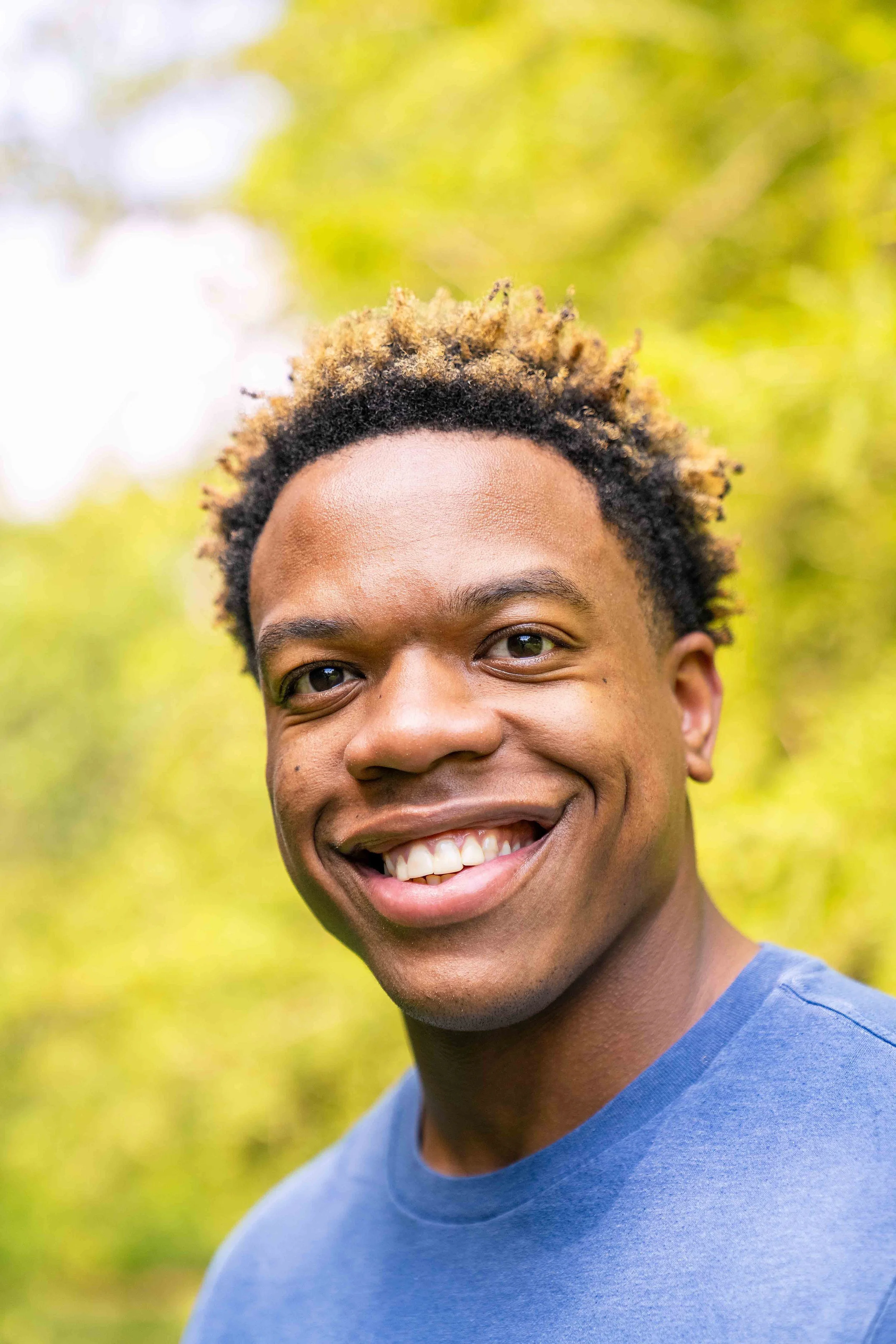 Close-up of a young man smiling outdoors, showing his teeth, with short curly hair and wearing a blue shirt against a blurred green background.