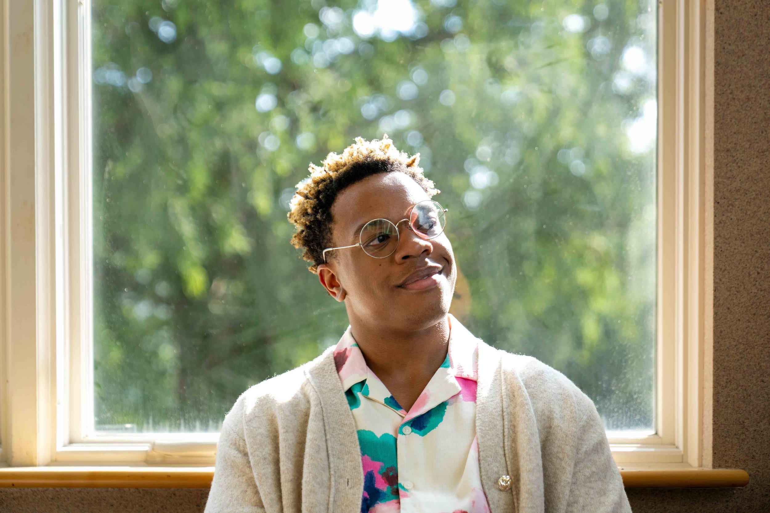 A person with short, curly, blonde-tipped hair wearing round glasses, a colorful shirt, and a cream cardigan, is standing in front of a window with green trees outside.