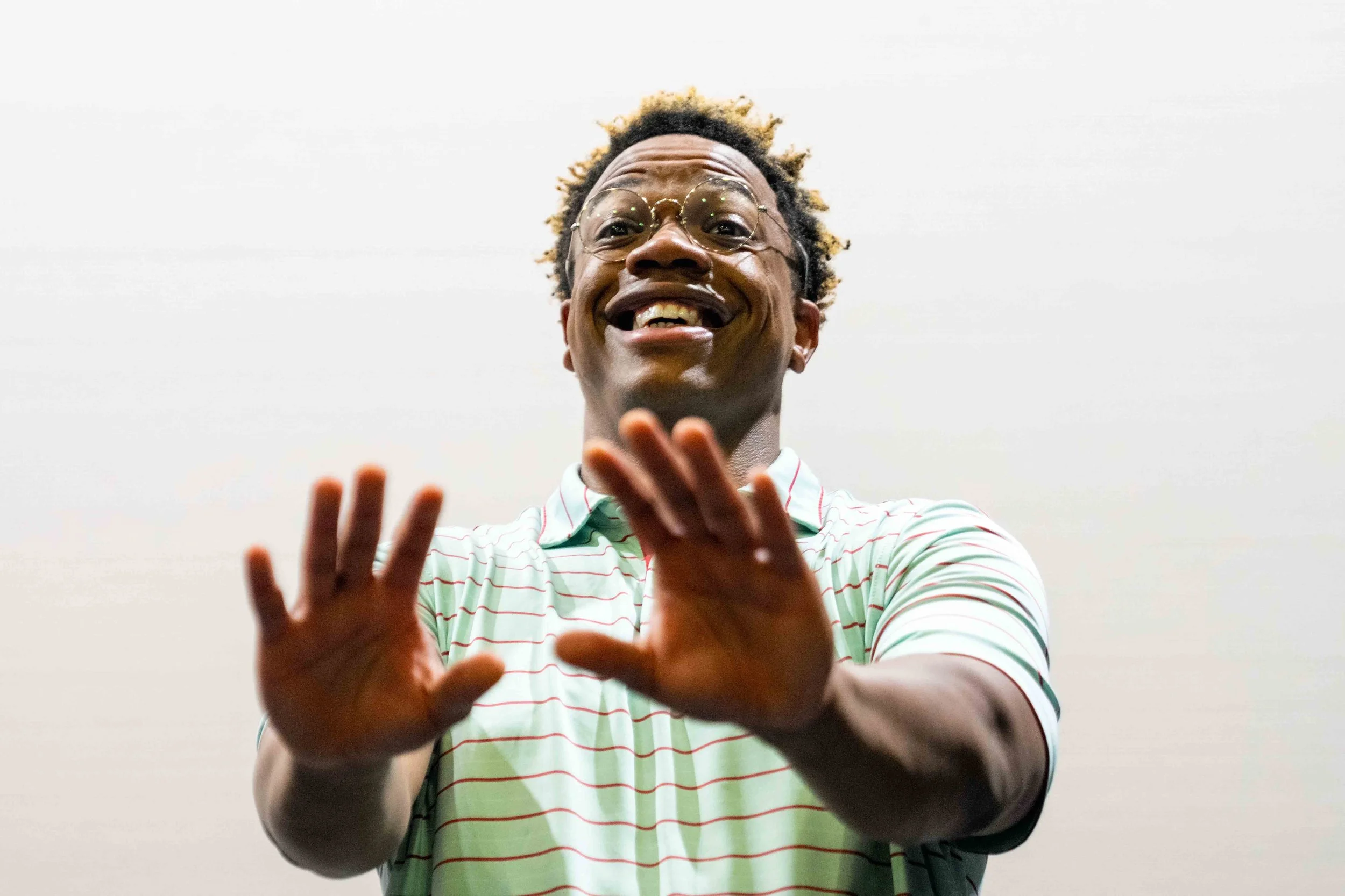 A man with curly hair and glasses smiling and reaching out with his hands in front of a plain white wall.