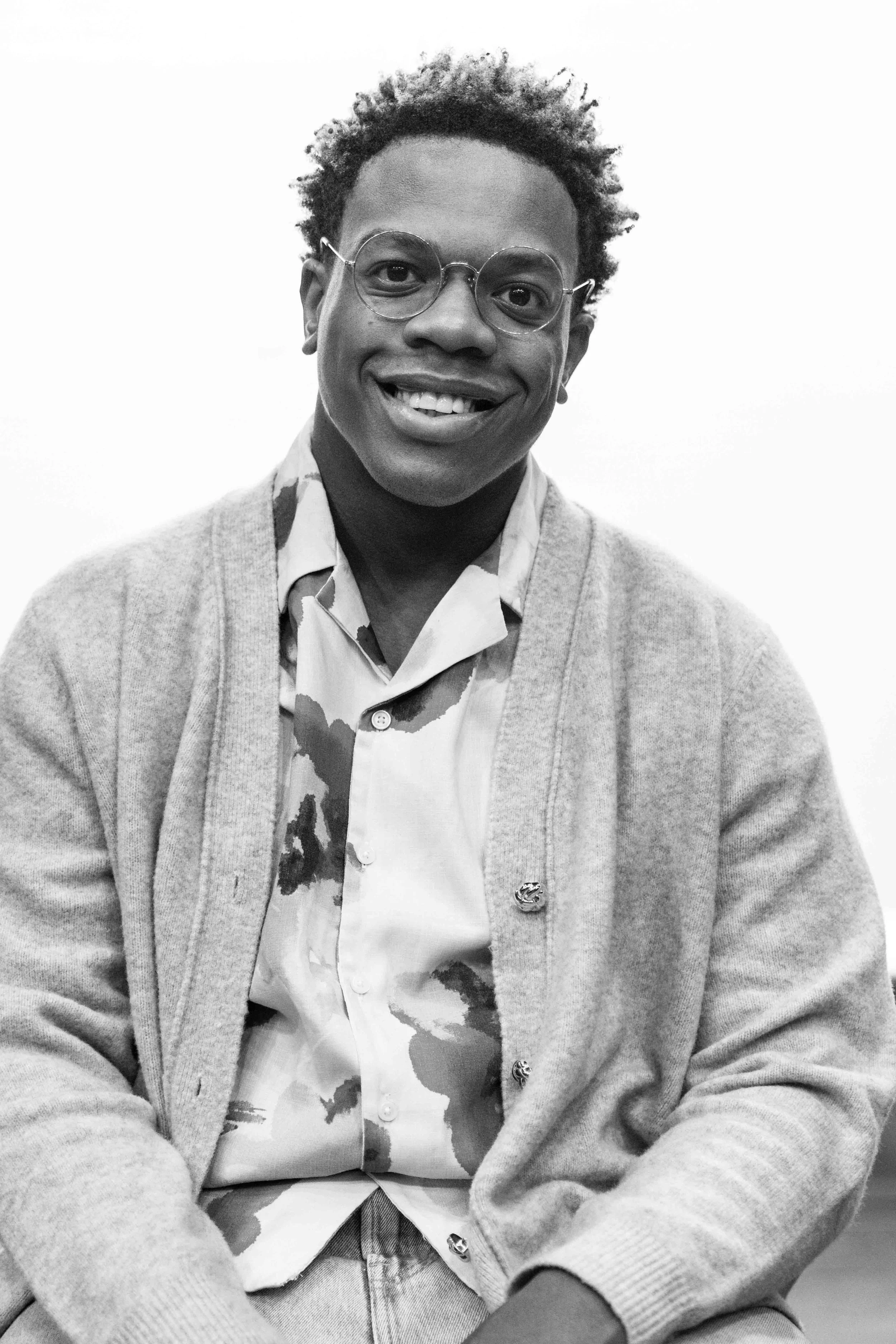 A young man with glasses, curly hair, and a warm smile, wearing a patterned shirt and a knitted cardigan, sitting with hands crossed, against a plain background.