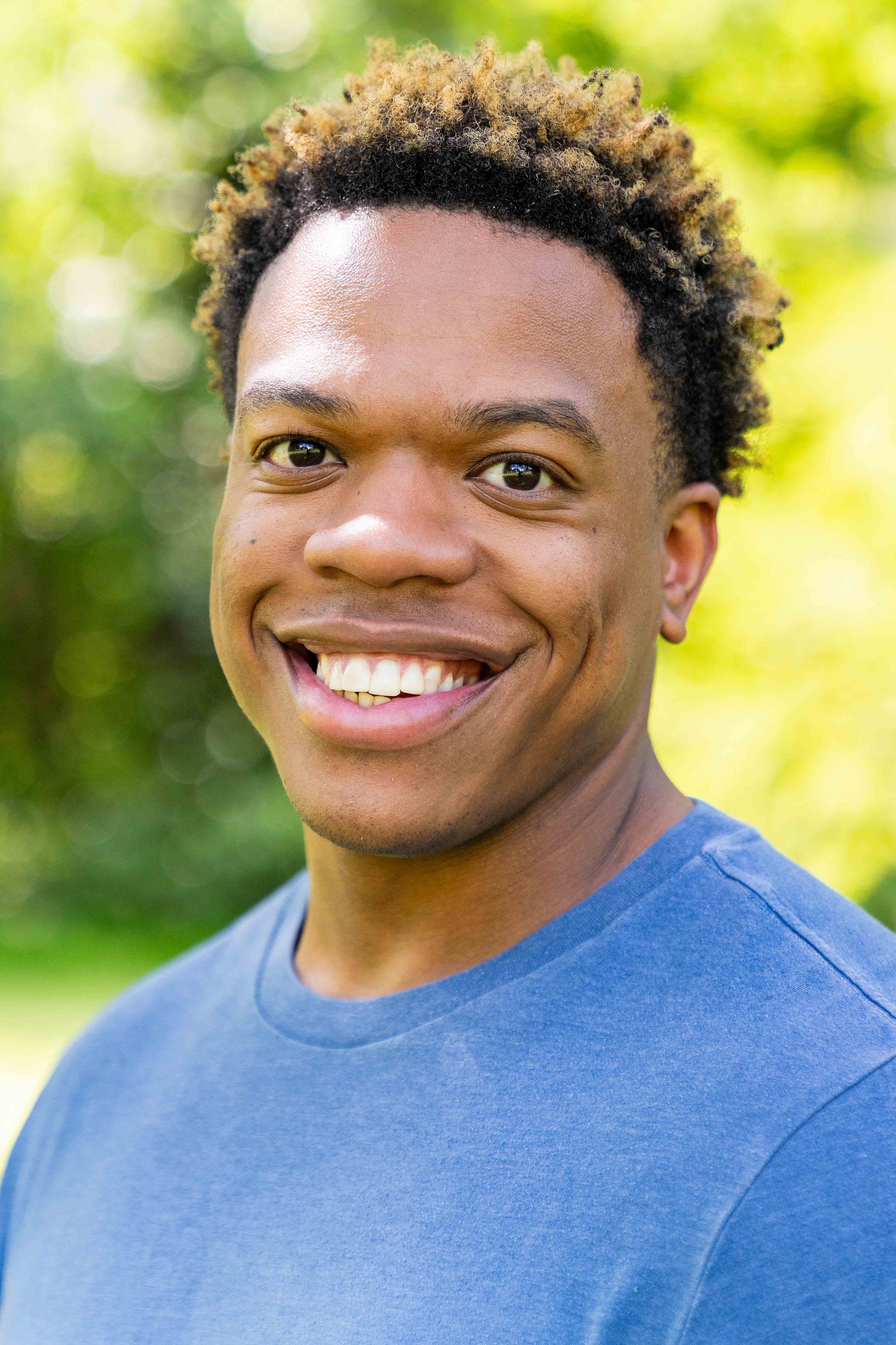 Close-up of a smiling young man with short curly hair, wearing a blue shirt, outdoors with a blurred green and yellow background.