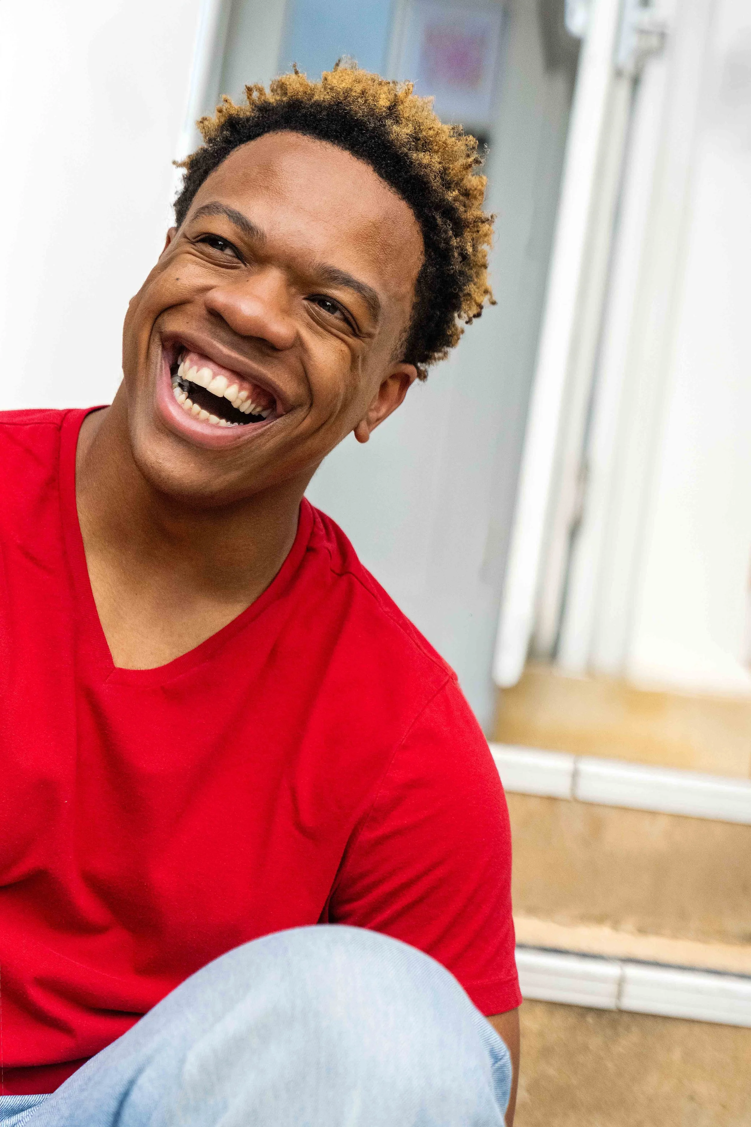 A young man with short, curly, two-tone hair smiling widely, showing his teeth. He is wearing a red shirt and appears to be indoors, near a white wall and door.
