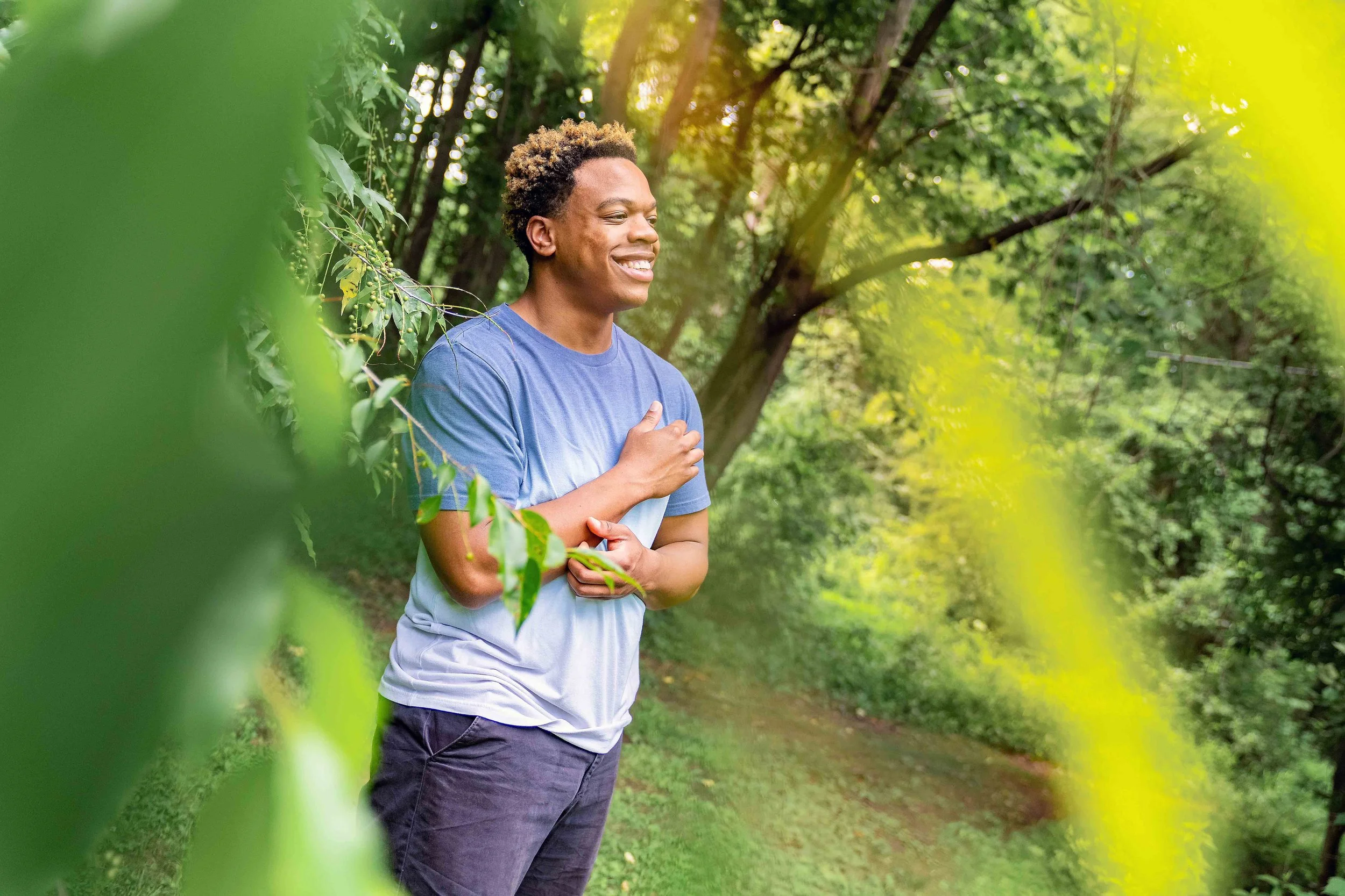 A young man smiling and standing among trees and greenery in a park, wearing a blue t-shirt and dark shorts, with sunlight filtering through the leaves.