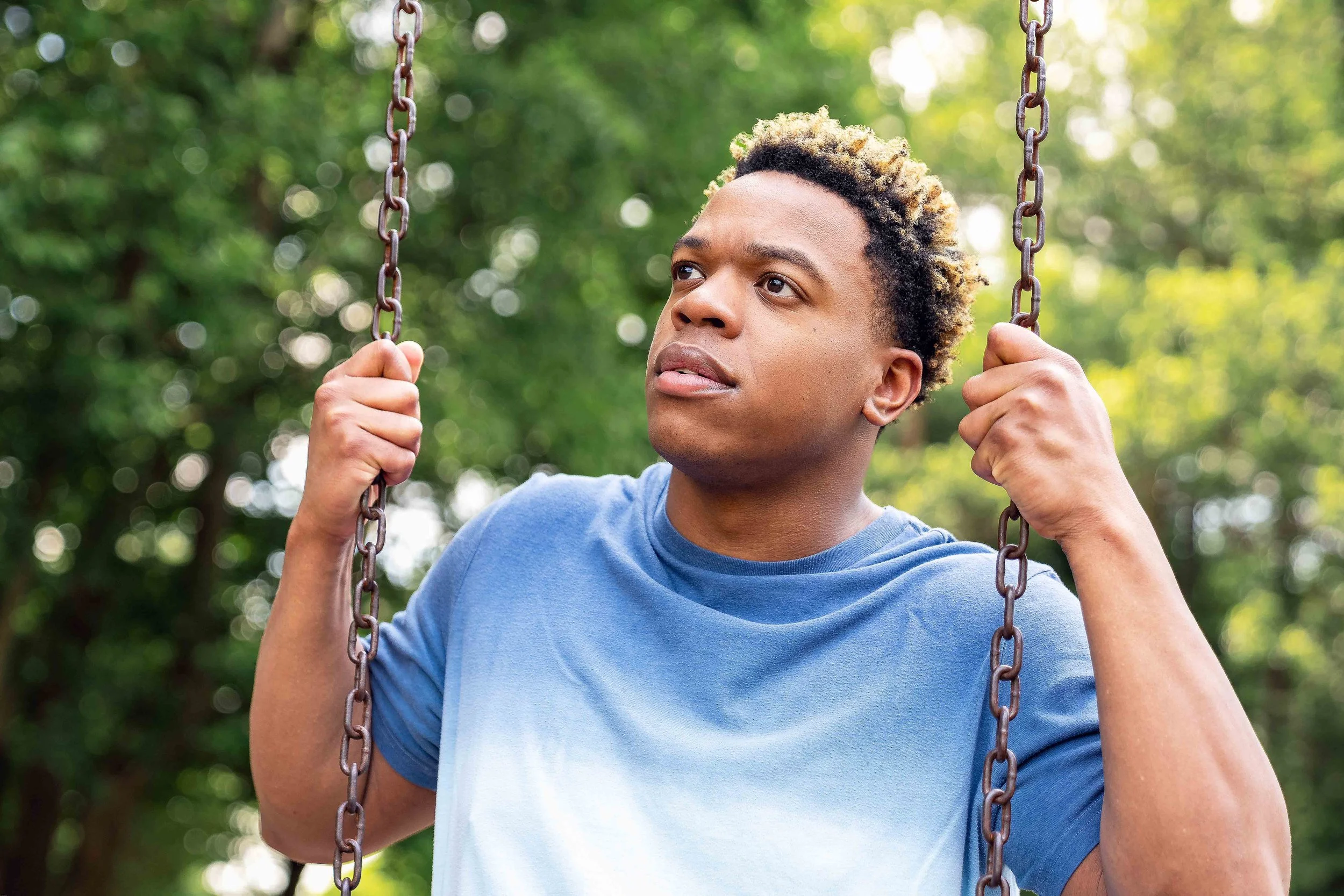 A young man with curly, highlighted hair wearing a blue t-shirt holding the chains of a swing outdoors surrounded by green trees.