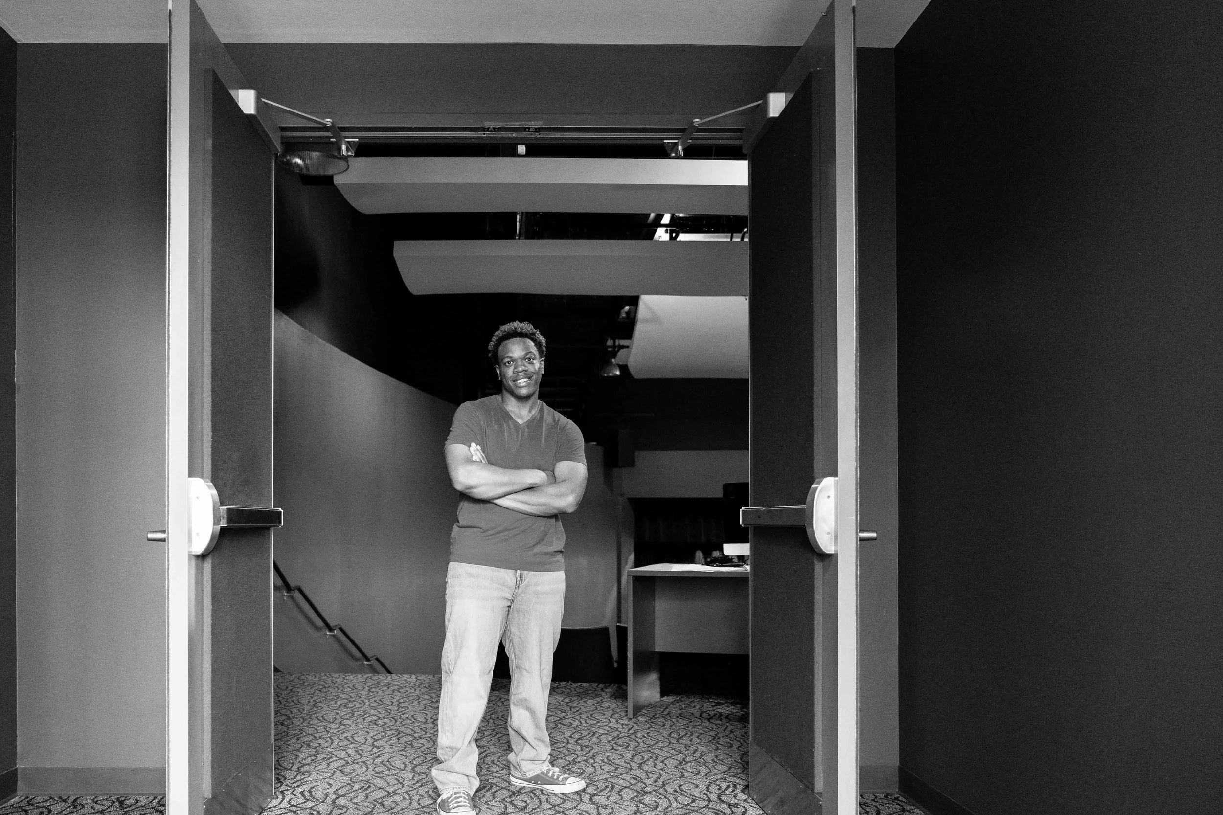 A young man with curly hair, wearing a T-shirt, jeans, and sneakers, smiling with arms crossed, standing in the doorway of an indoor space, with stairs visible in the background.