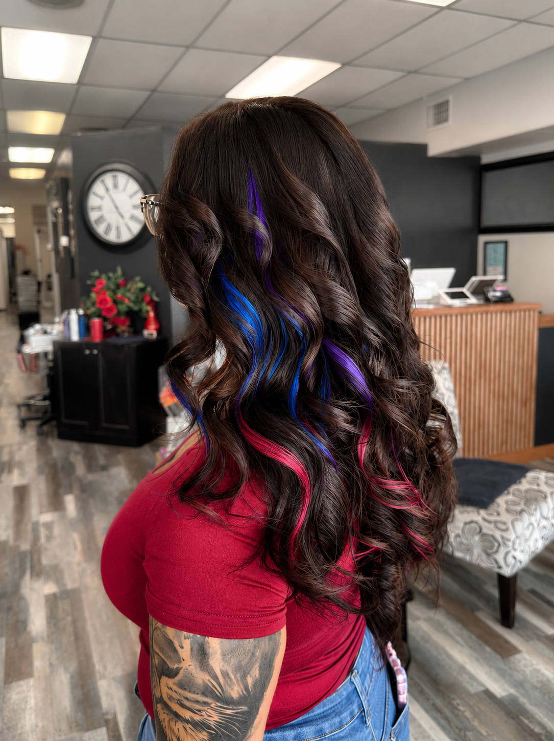 A woman with long, dark, wavy hair with blue, purple, and pink streaks, wearing glasses and a red shirt, standing in a hair salon.