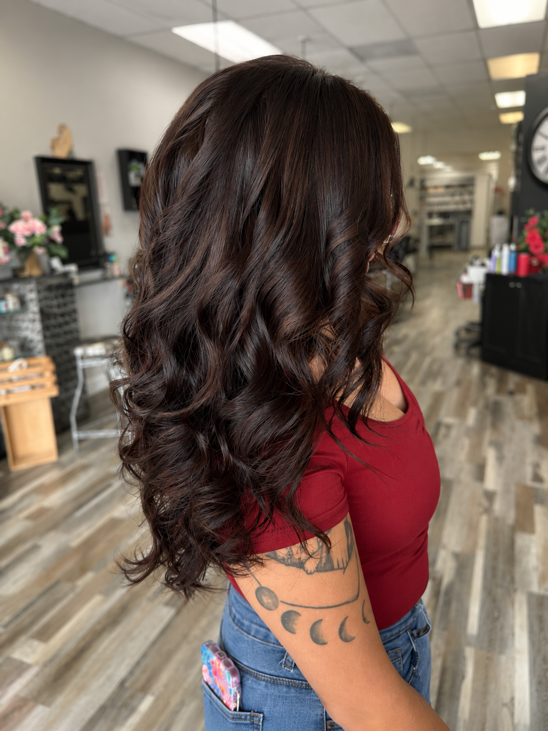 A woman with long, dark, wavy hair wearing a red t-shirt and blue jeans, standing in a salon.