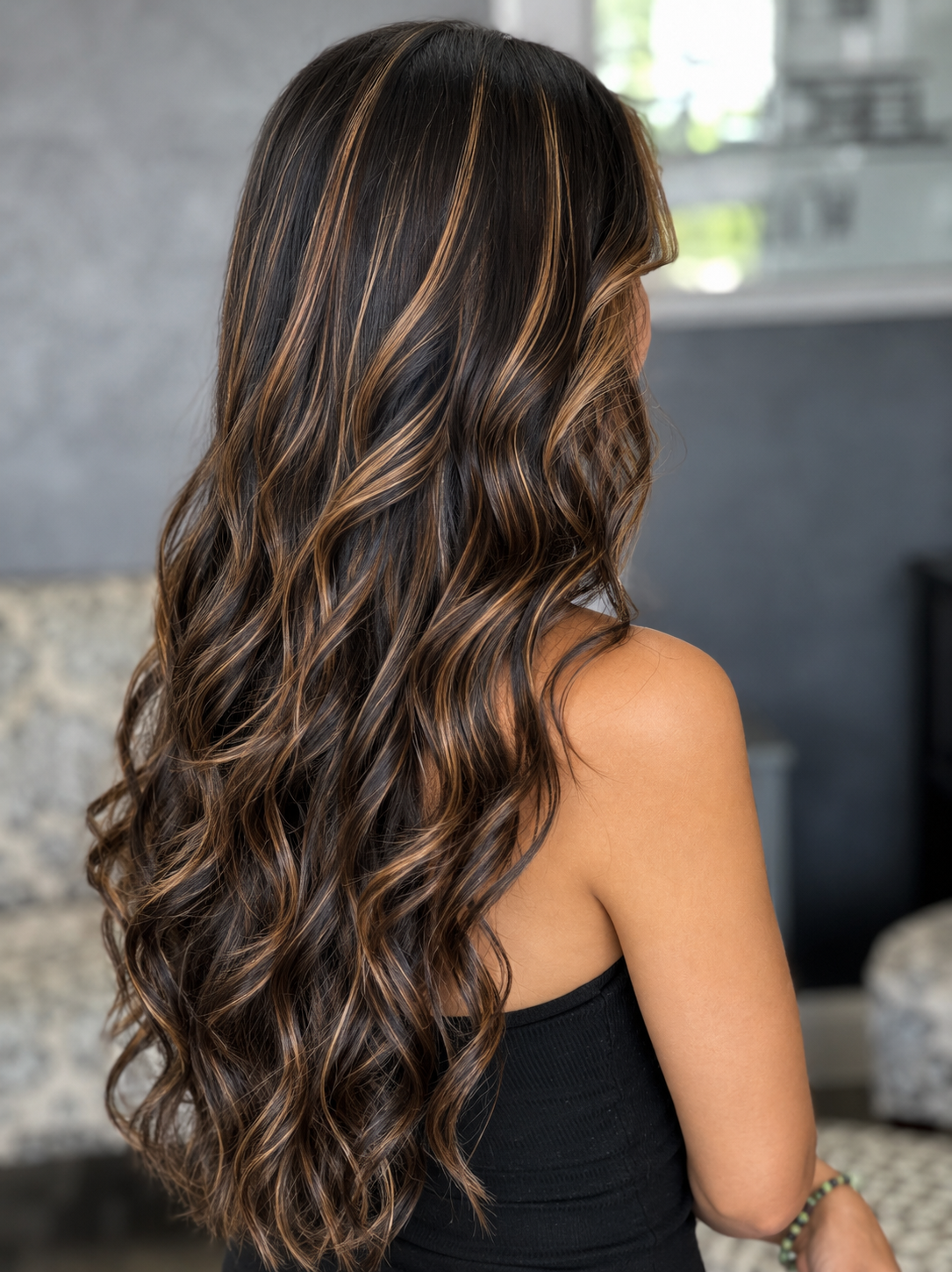 Back view of a woman with long, wavy hair featuring caramel highlights, wearing a black sleeveless top, in a modern indoor setting.