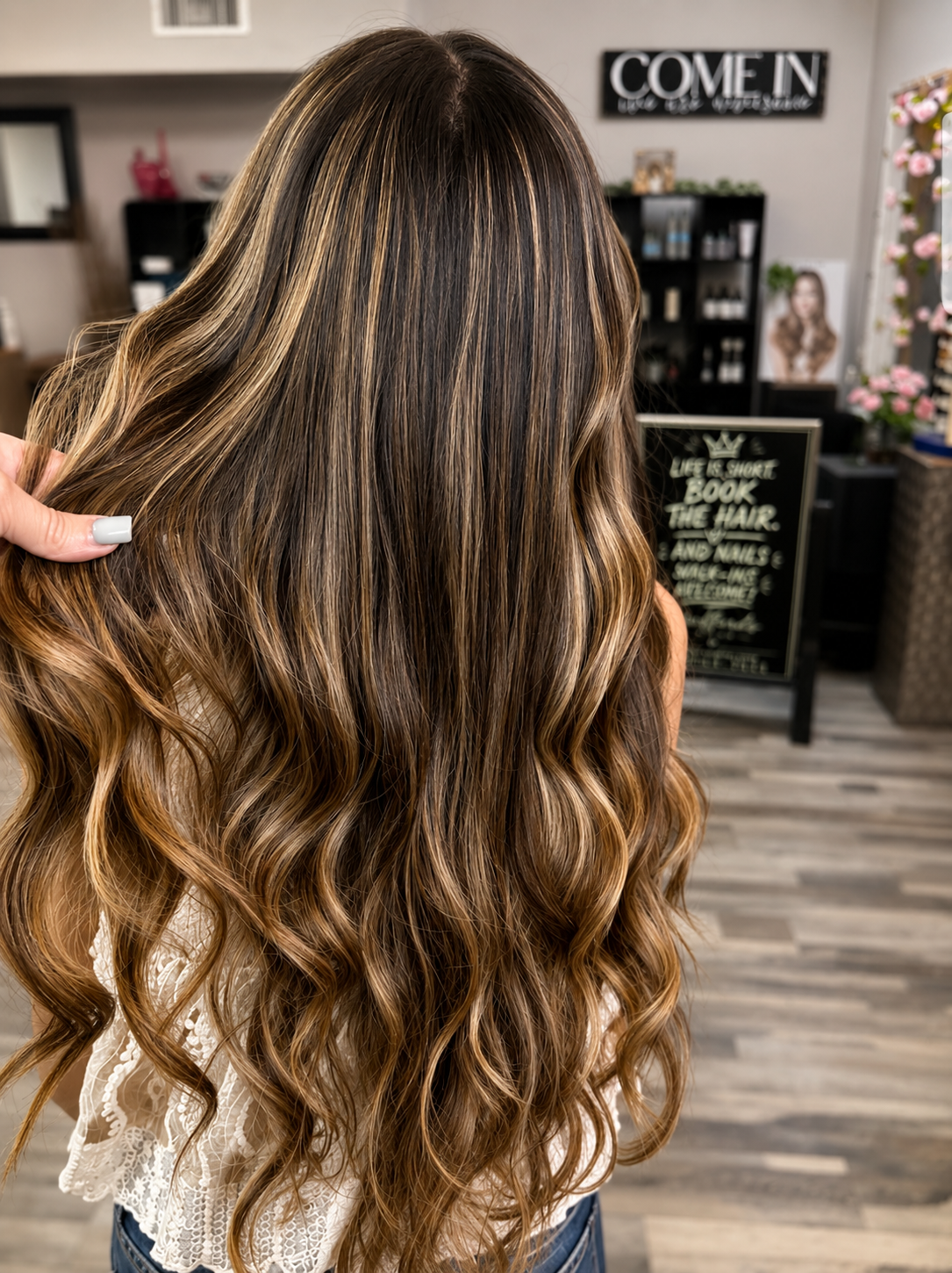 A woman with long, wavy, caramel-colored hair with highlights inside a salon.