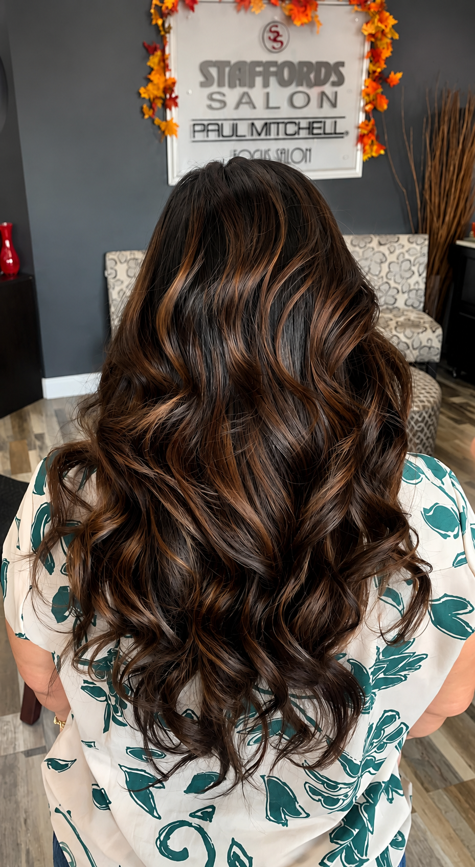 Back view of a woman with long, wavy hair dyed with brown and caramel highlights, sitting in a salon with a sign and chairs in the background.