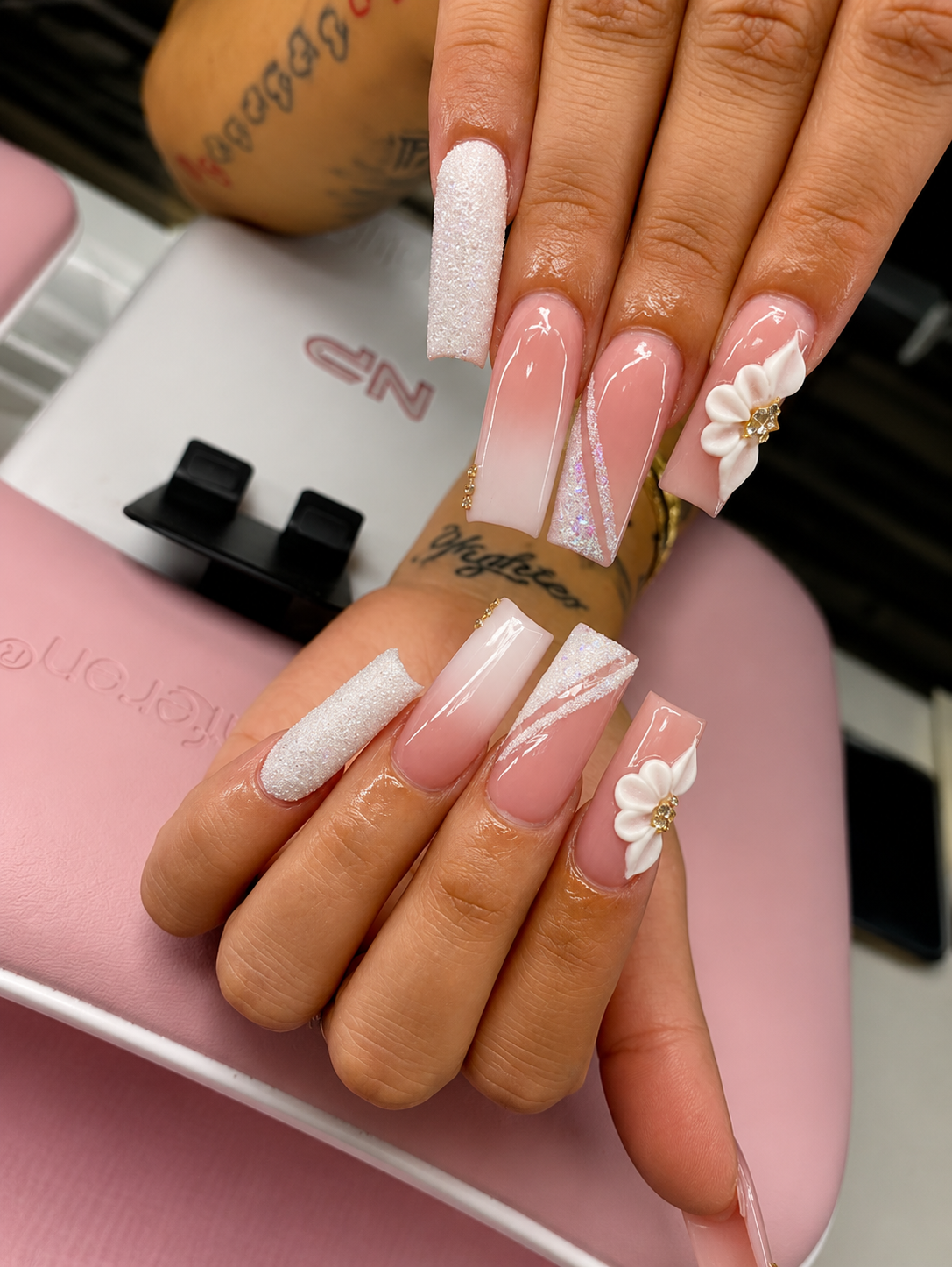 Close-up of a woman's hand with long, elegant, pink and white gradient acrylic nails, decorated with 3D floral accents and glitter, resting on a pink surface near a nail dryer.