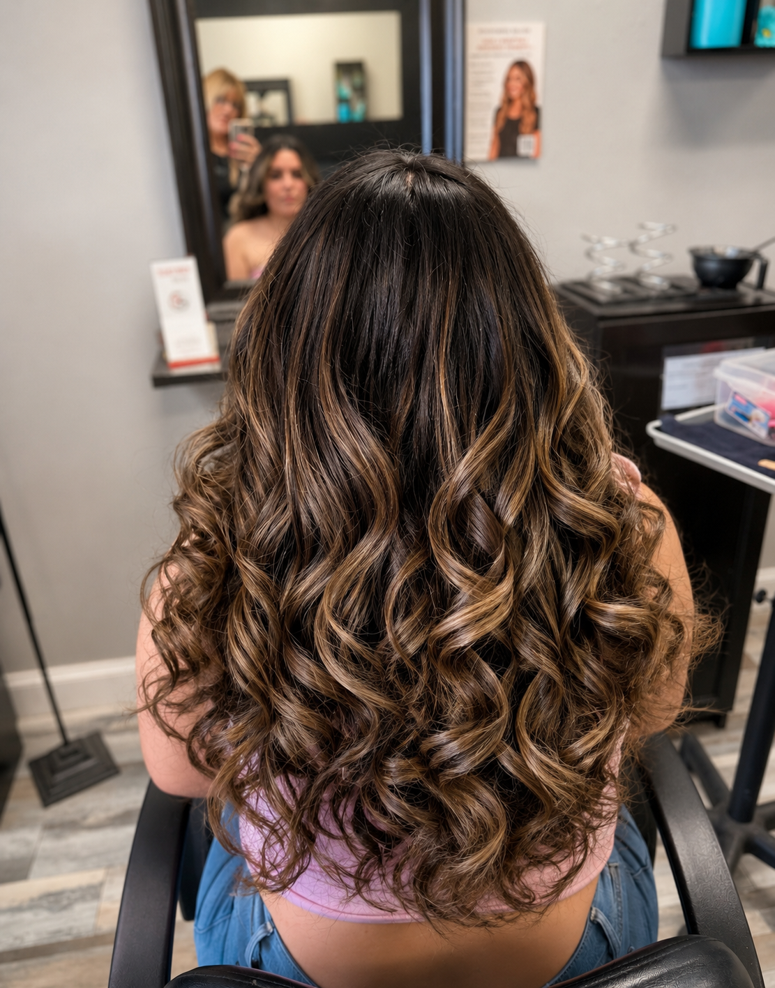 Back of a woman with long, brown, curled hair sitting in a salon chair, facing a mirror.