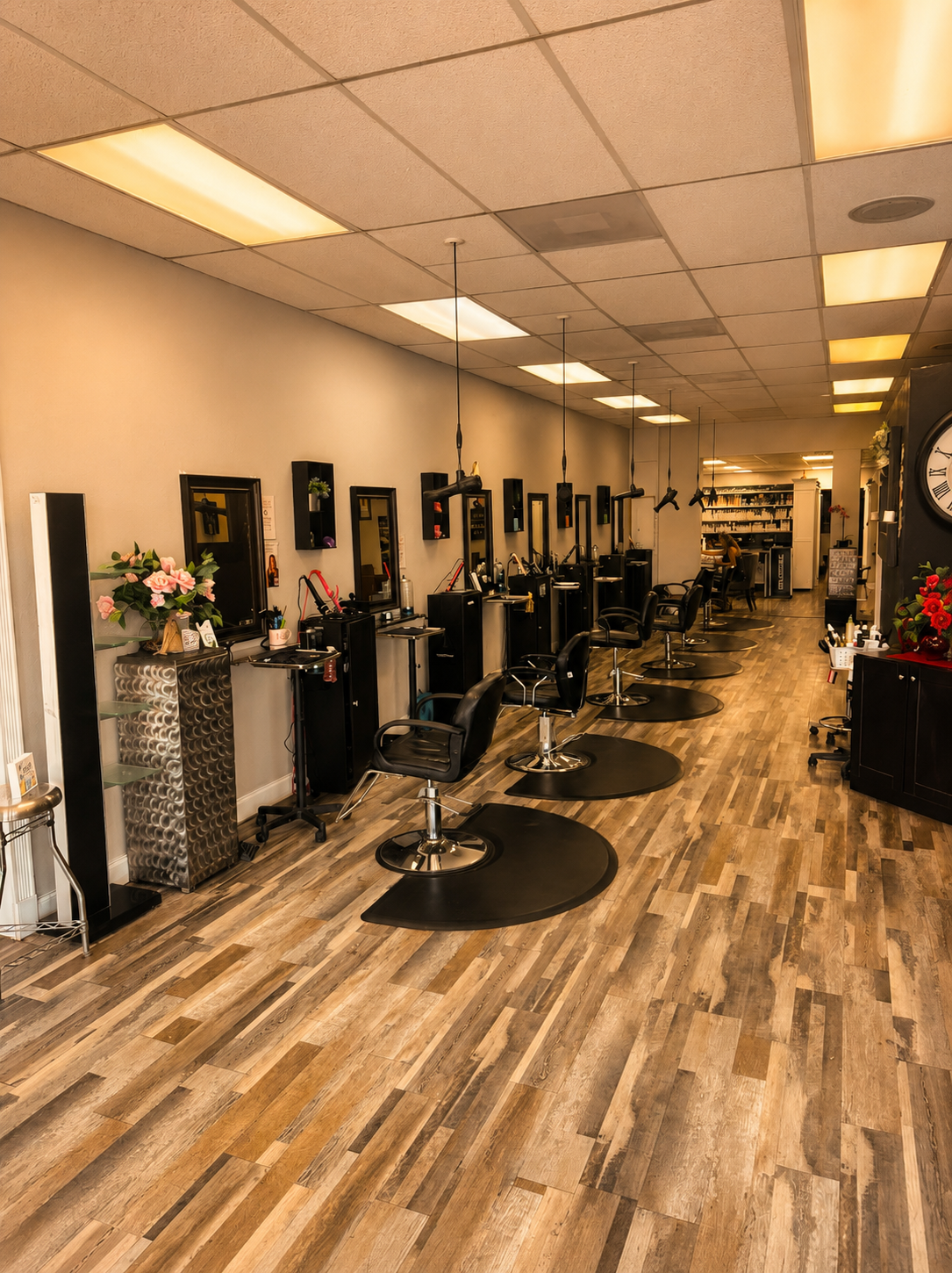 Empty hair salon with multiple black salon chairs, styling stations with mirrors, wooden flooring, and overhead lighting.