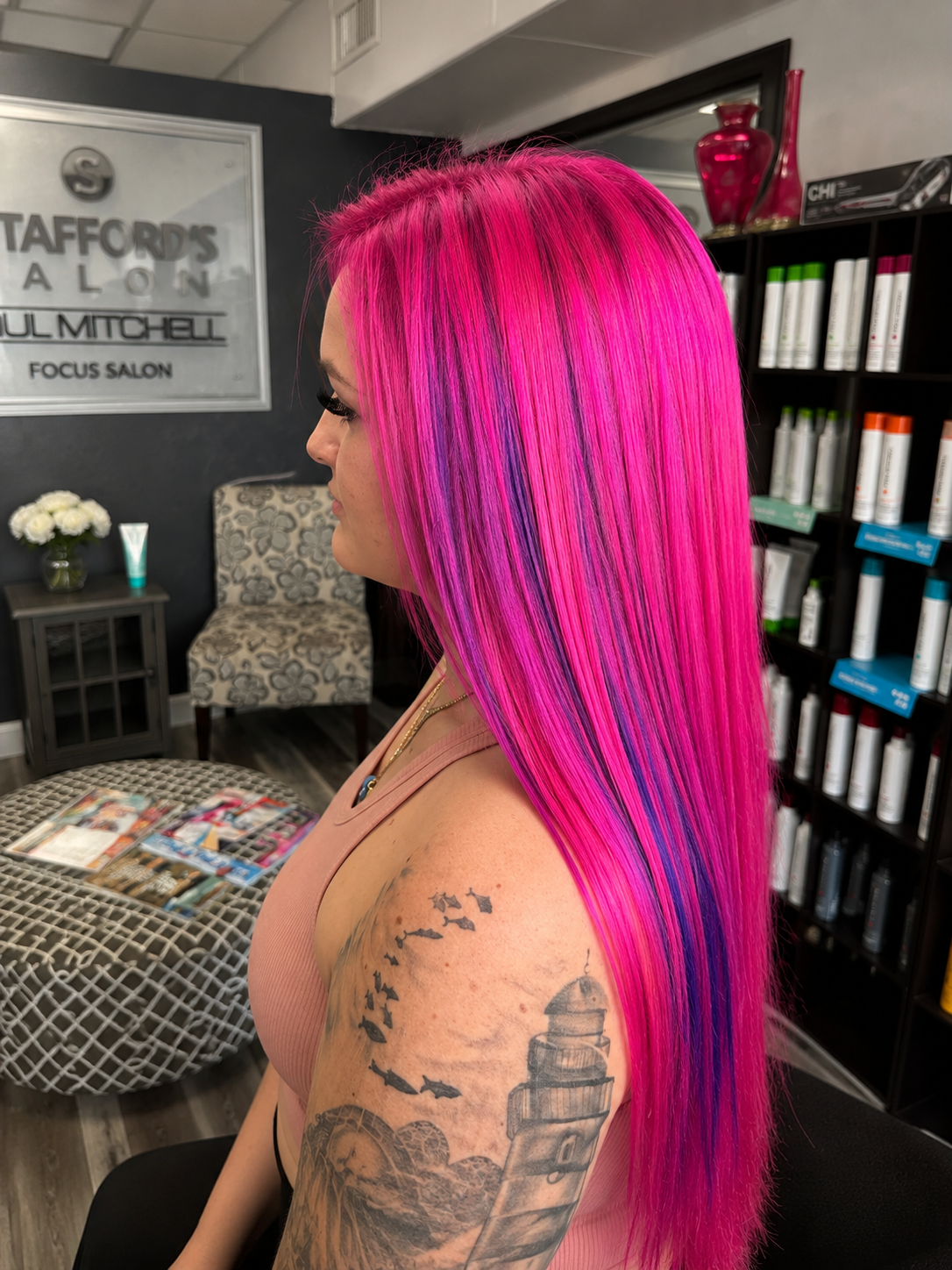 A woman with vibrant pink and purple straight hair sitting in a salon, showing her tattoos of a lighthouse and birds on her arm, with salon shelves and a seating area in the background.
