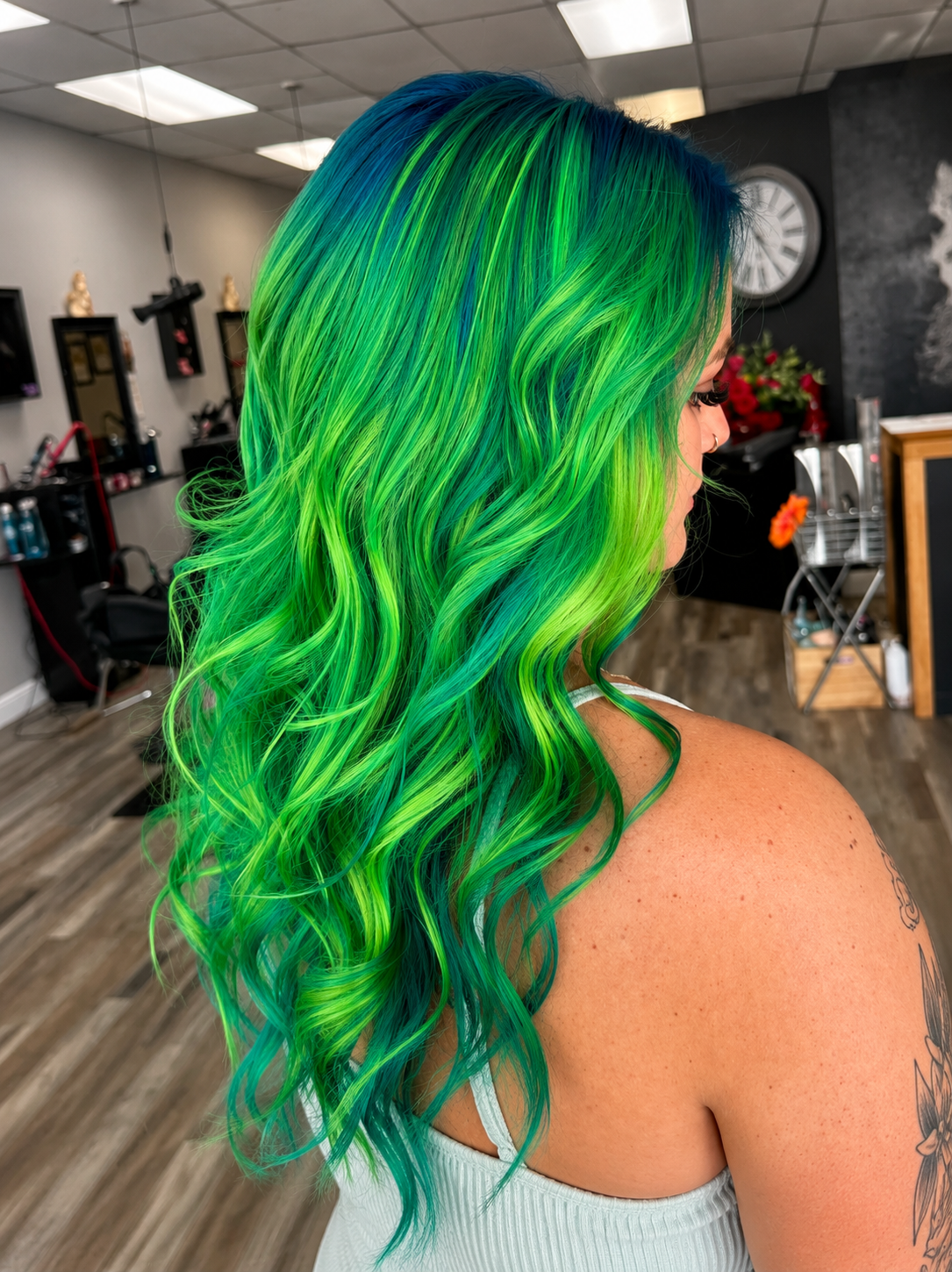 Woman with vibrant rainbow-colored hair in shades of green, blue, and yellow, styled in loose waves, inside a hair salon.