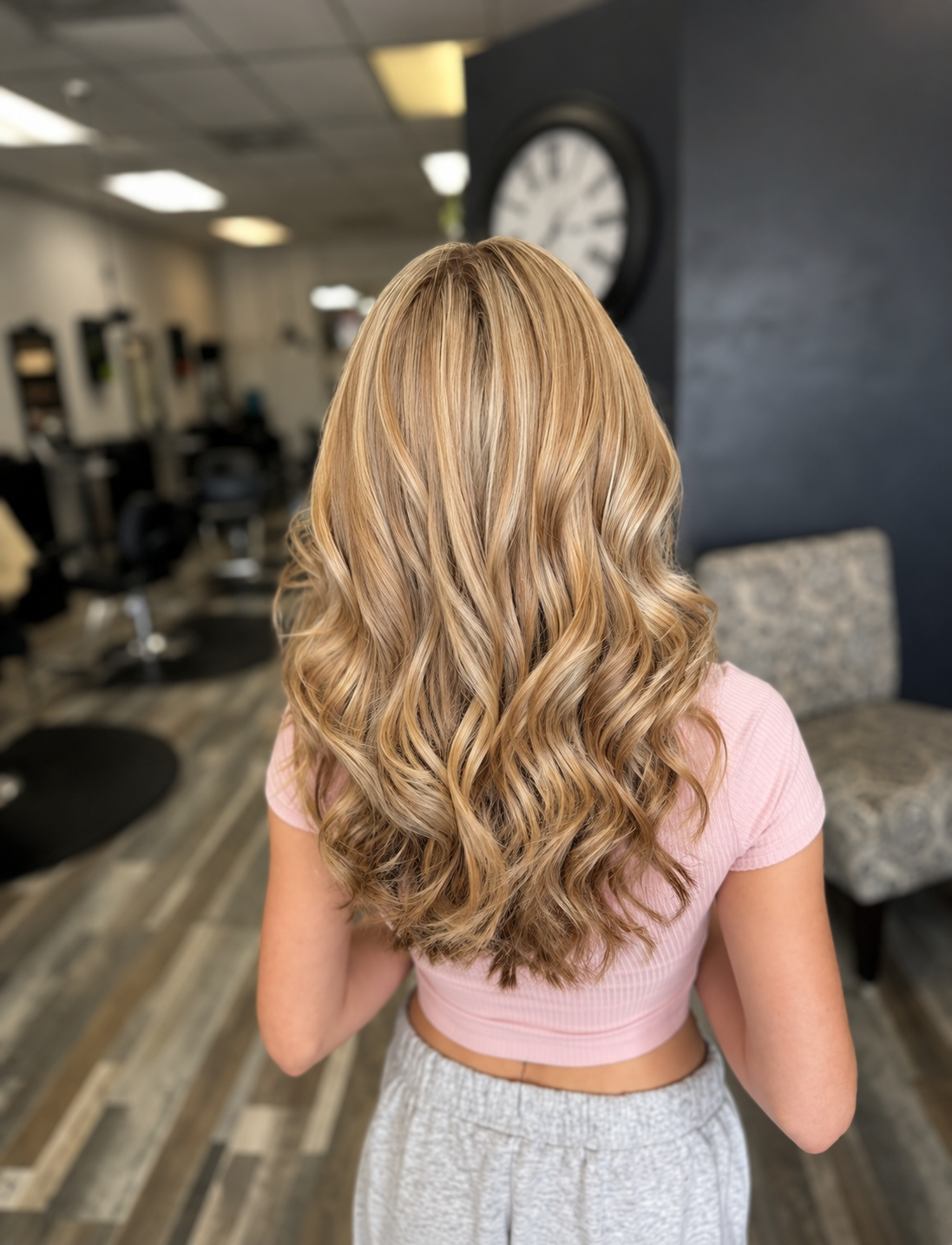 Back view of a woman with long, wavy blond hair sitting in a hair salon.